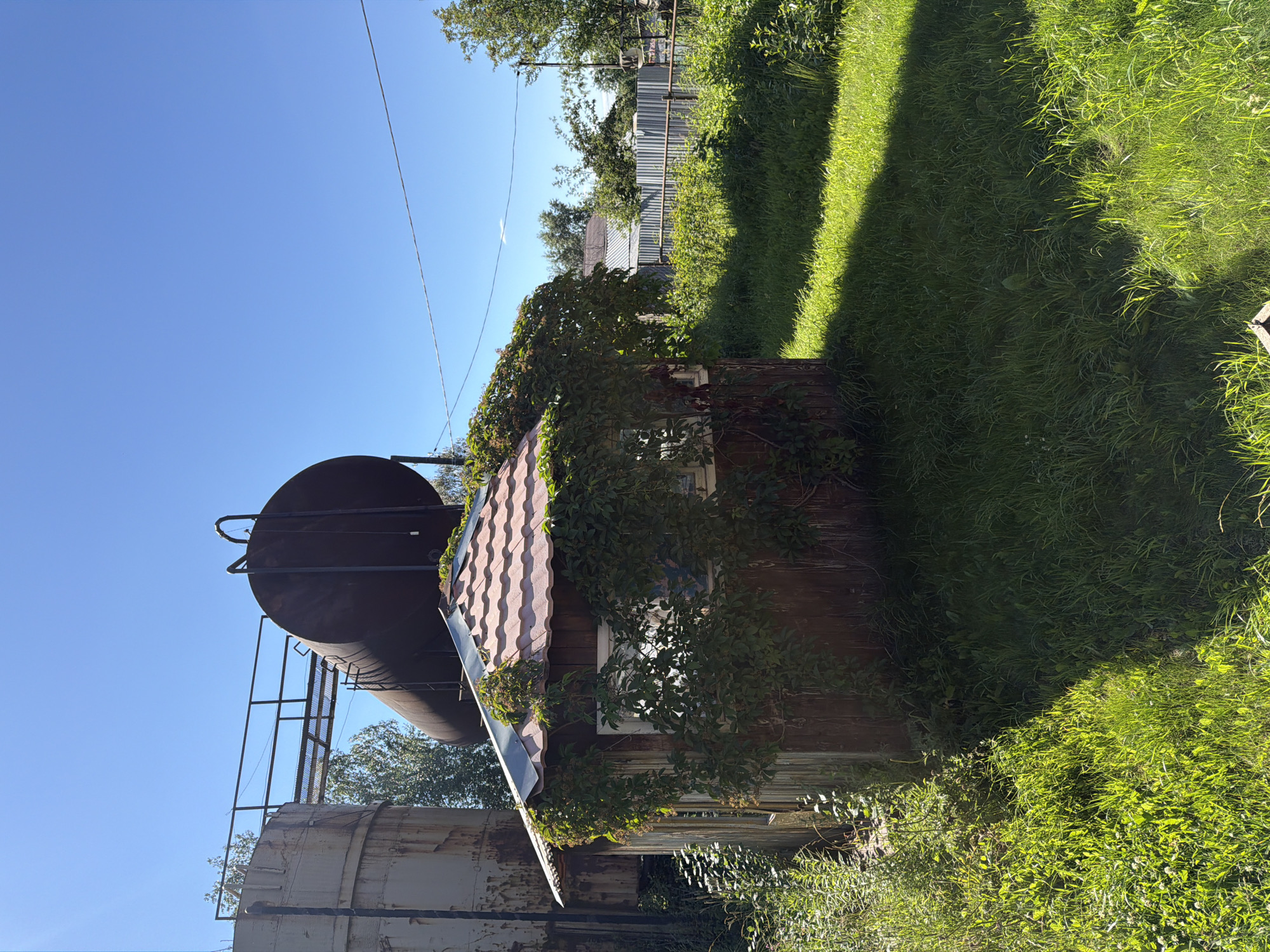 The image depicts an outdoor scene on a clear day. In the foreground, a small, dilapidated wooden shed with a red tile roof is covered in green ivy. A large, rusted cylindrical tank is positioned behind the shed, supported by metal structures. To the left of the shed, another large, weathered cylindrical structure is visible. The ground is covered in green grass, with a distinct shadow cast across it. In the background, a metal fence and trees are present. The scene appears to be located in an industrial or semi-industrial area, possibly on the outskirts of Plamya, Russia, given the presence of large tanks and industrial-looking structures, contrasting with the more natural elements of grass, ivy, and trees. There are no people or visible actions taking place in the image.