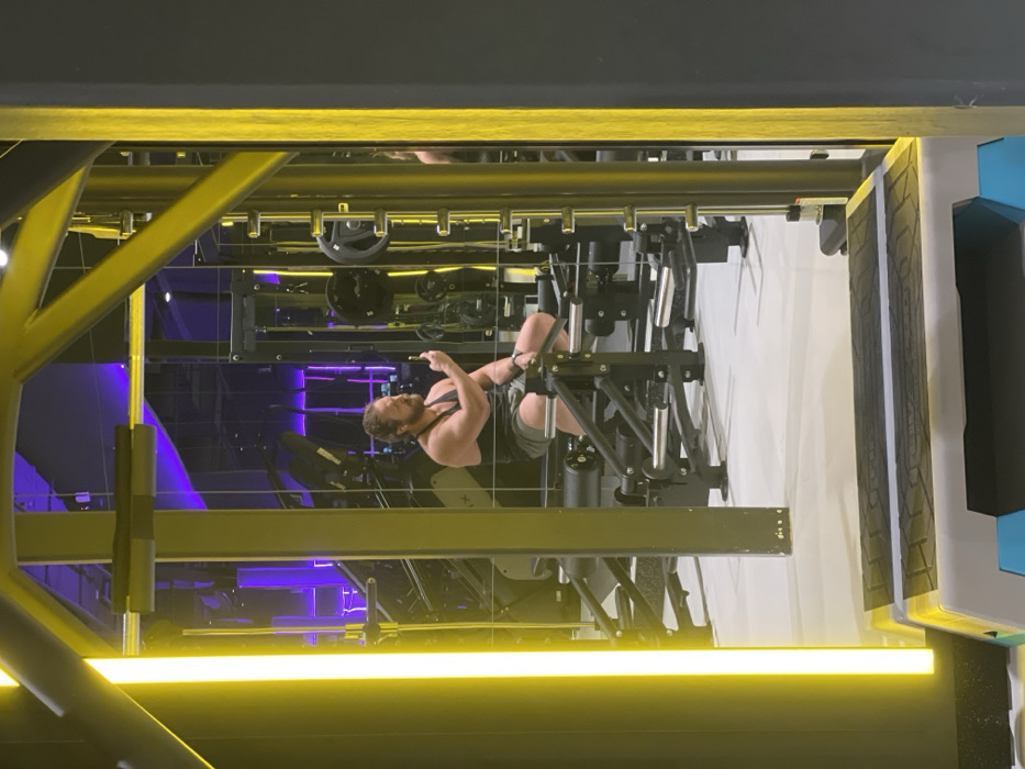 This image depicts a muscular man using gym equipment within a modern fitness facility in Saint Petersburg, Russia. The man, with a beard and curly hair, is wearing a dark tank top and dark shorts. He is positioned on a weight machine, possibly a leg press, and appears to be looking down at his hands, potentially interacting with a phone or adjusting the apparatus.
The setting is characterized by its contemporary design, featuring striking linear yellow lights that frame the bottom and top of the visible area. The background is highly reflective, likely consisting of large mirrors or polished surfaces, which create a layered visual effect, showing reflections of other gym equipment and ambient purple lighting from deeper within the facility. Various black weight plates are visible on the machinery. A structural element with a blue accent can be seen on the far right. The scene is illuminated by artificial lighting, contributing to a bright and dynamic atmosphere. No discernible text is visible.