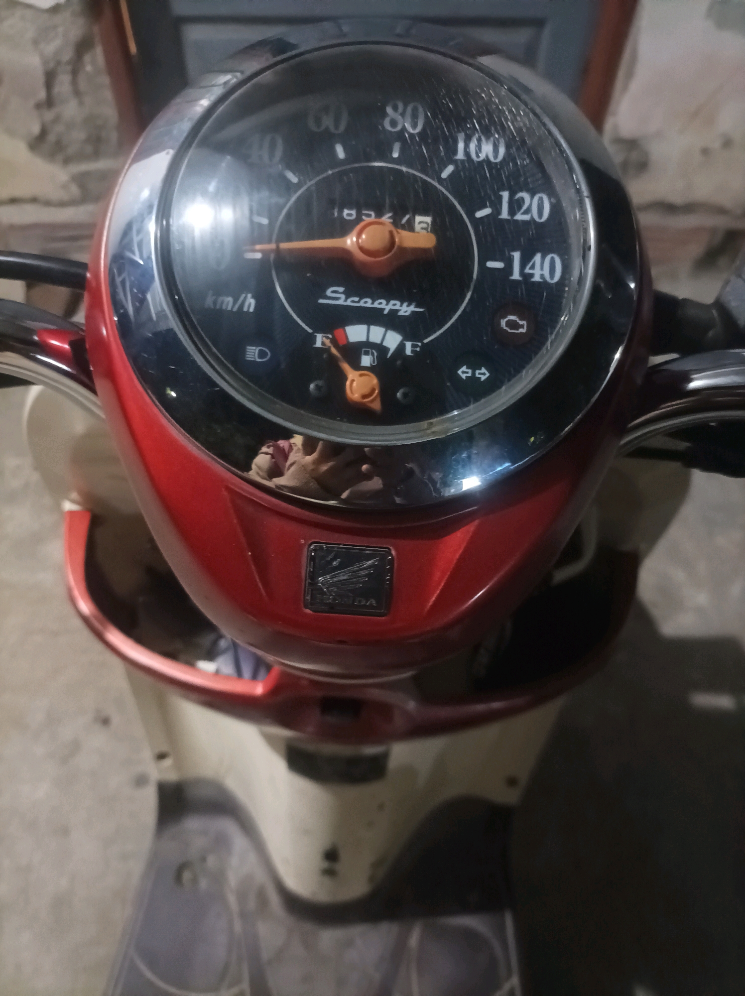 The image shows the dashboard of a red and cream-colored Honda Scoopy motorcycle. The speedometer displays a speed of approximately 40 km/h, and the fuel gauge indicates the tank is nearly full. The engine warning light and turn signal indicators are visible. A reflection of a person's face and arm holding the camera is visible on the curved surface of the dashboard. The motorcycle is parked on a concrete surface, likely outdoors or in a garage, with a stone wall in the background. The scene is located in Canga'an, Indonesia.