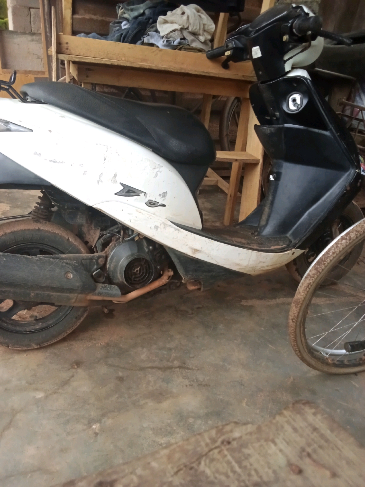 A dirt-covered white and black scooter is parked on a concrete floor, in what appears to be an open-sided shelter or shed. The scooter has a black seat, a white front fairing, and a black rear section. The engine is visible and appears to be exposed to the elements, with dust and dirt accumulating on it. A portion of a bicycle wheel is visible to the right of the scooter. In the background, a wooden shelf holds a pile of various textiles, possibly clothing. The overall environment is one of disarray and neglect, with dirt and dust pervasive throughout the scene. The scooter's condition suggests it may have been stored outdoors or in a dusty environment for an extended period.