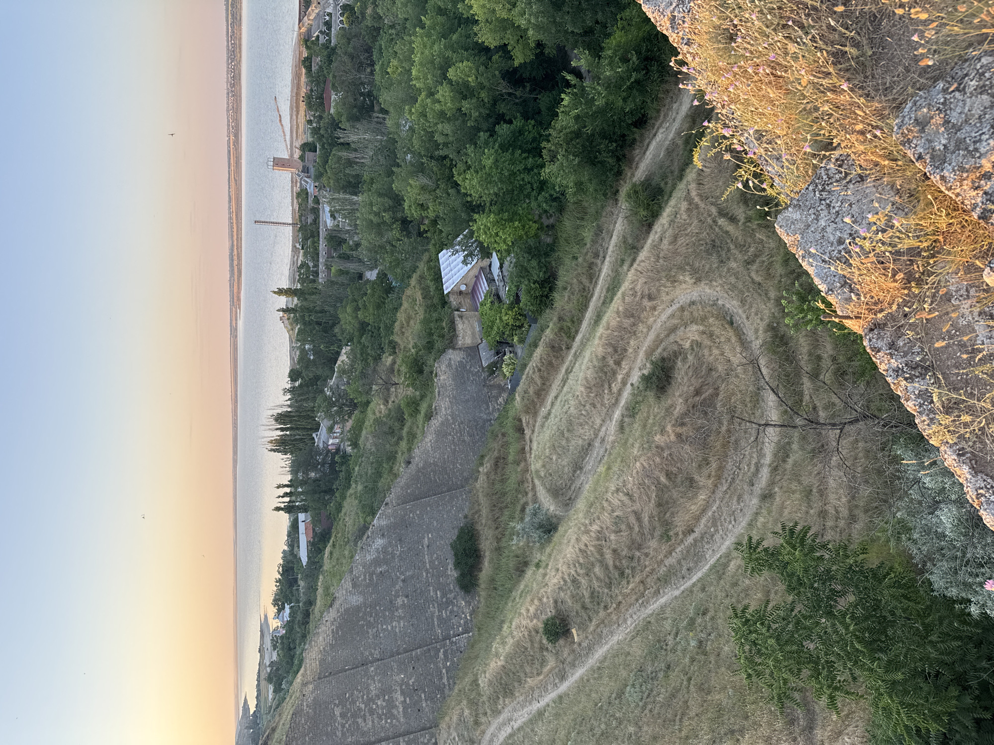 The image depicts a panoramic view overlooking a body of water and a settlement at sunset. In the foreground, a steep, rocky incline covered with dry grass and small purple flowers is visible. Below this, a winding dirt path cuts through a dry, grassy hillside. Further down, a cluster of trees and buildings, including a house with a white roof, are nestled amongst lush green foliage. Across the water, a distant shoreline is lined with buildings and industrial structures, including a tall tower with a light and a smaller tower with a red top. The sky displays a soft gradient of orange and blue, indicative of either sunrise or sunset. The scene appears to be the coastal area of Mizhlymanske, Ukraine.