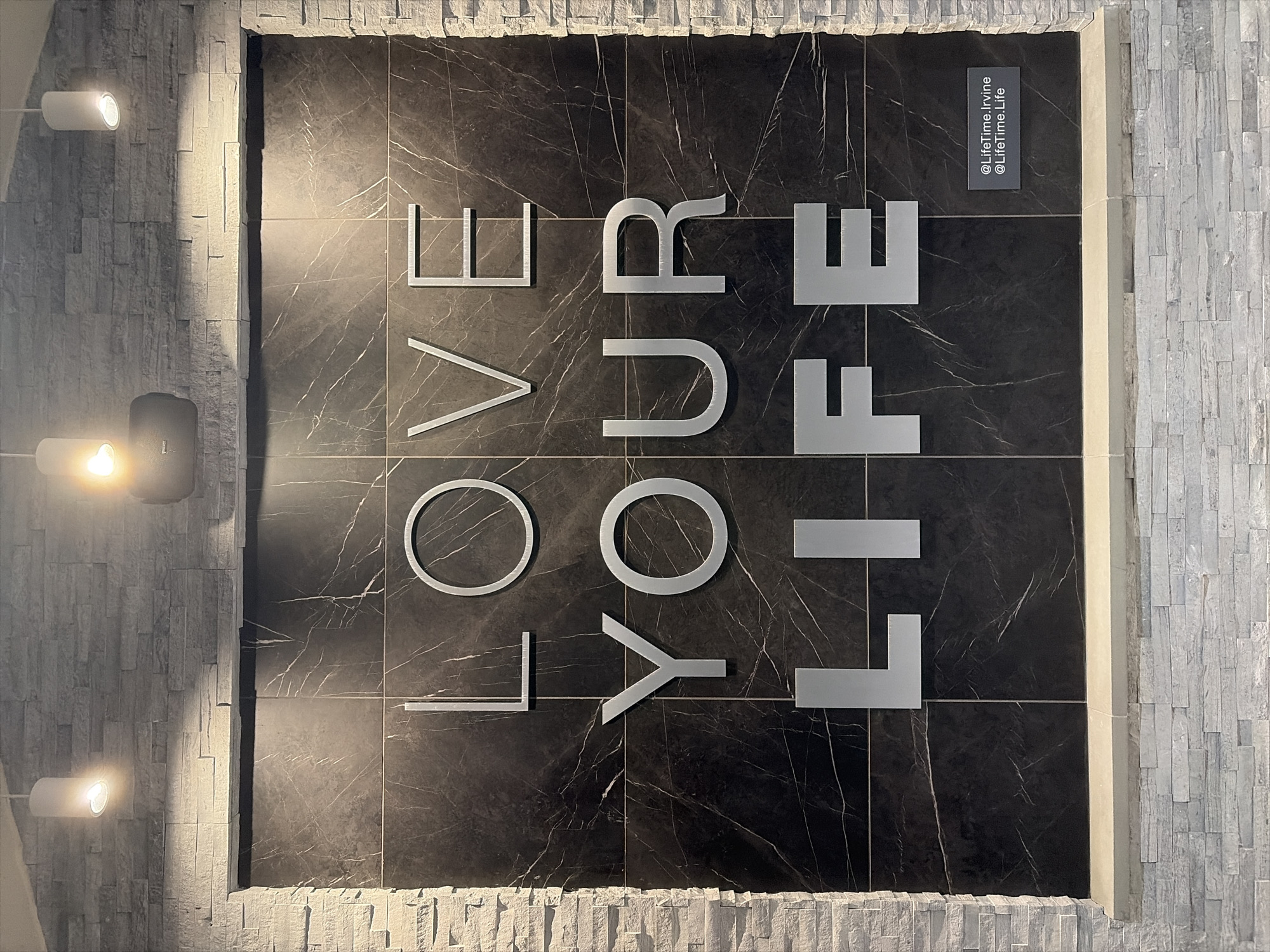 Here is a detailed description of the image:
The image shows a decorative wall display with the words "LOVE YOUR LIFE" arranged vertically on a dark, marble-patterned tile surface. The letters are large, sans-serif, and appear to be made of a metallic material. To the right of the word "LIFE" is a small, vertical plaque or sign that reads "@LifeTime.Irvine @LifeTime.Life". The tile surface is framed by a border of textured, light-colored stone or brick. On the left side of the frame, three wall-mounted spotlights are visible. Based on the sign, the location is likely a Life Time fitness center in Irvine, California, United States of America. The scene is well-lit, with the spotlights enhancing the visual impact of the display. No people or direct actions are visible. The primary interaction is the viewer's engagement with the motivational message and branding of the Life Time facility.