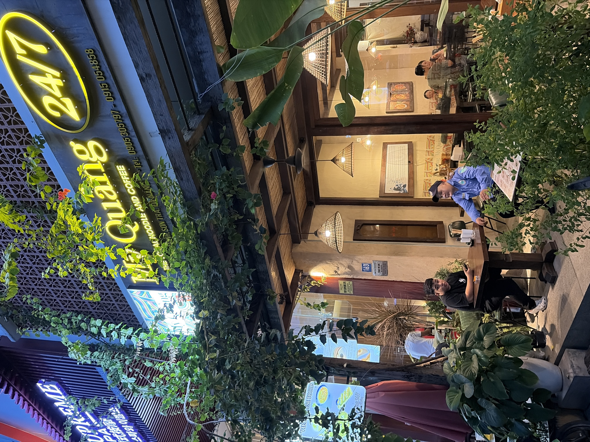 This is a nighttime outdoor shot of a restaurant called "My Quang" in Da Nang, Vietnam. The restaurant sign, illuminated in bright yellow neon, reads "My Quang" and "24/7". Below the main sign, there is smaller text indicating "THE LOCAL NOODLE AND COFFEE" and phone numbers. The restaurant appears to be a street-facing establishment with large windows, allowing a view into the interior where several patrons are seated. Some of the patrons are visible at tables, while others are obscured. The exterior features lush green foliage and potted plants, contributing to a vibrant and inviting atmosphere. A single neon sign on an adjacent building, partially obscured by foliage, reads "SUPREME" in blue lettering. The overall scene depicts a lively dining spot in a Vietnamese city.