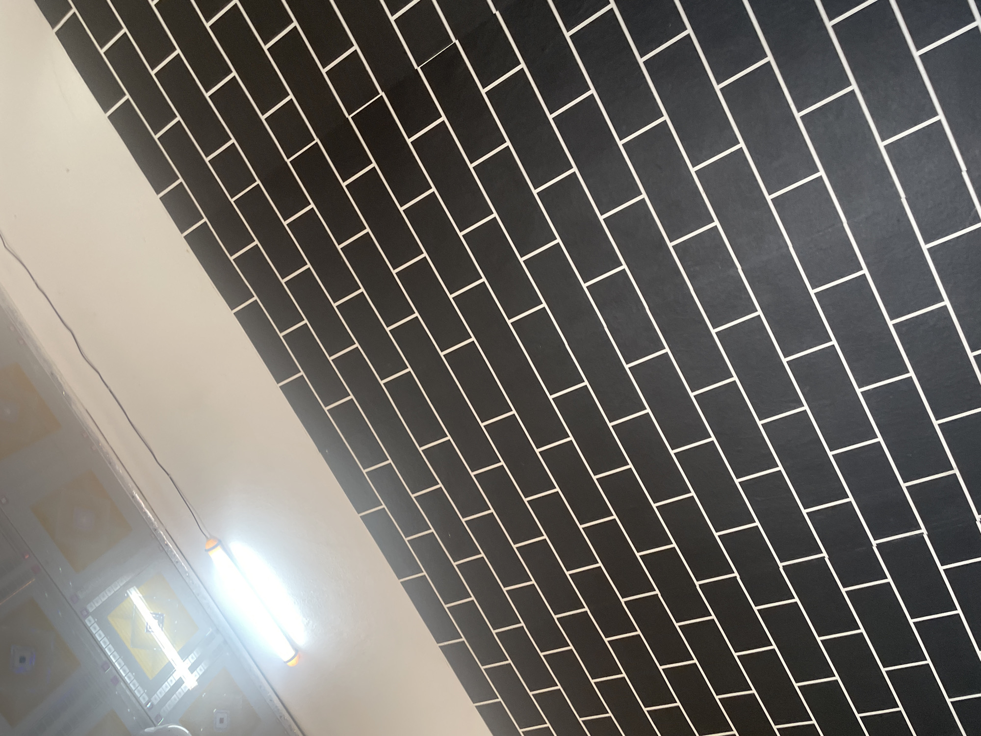This media file shows a view of a ceiling or a wall covered in black rectangular tiles with thin white grout lines, arranged in a staggered pattern. The tiled surface is diagonally oriented within the frame, filling the right and upper portions. To the left of the tiled area, there is a plain white surface, possibly a wall or ceiling. Hanging from the white surface is a fluorescent light fixture, which is brightly illuminated and appears to be a tube light with orange caps on its ends. A black wire connects the light to something out of frame. The lower left portion of the image shows some reflective surfaces and what appears to be a suspended ceiling with a pattern of yellow and white squares. The overall lighting suggests an indoor setting, likely during the day due to the bright illumination of the artificial light. There is no text visible in the image. The scene does not contain any people or animals. The mood is neutral and functional, focusing on the architectural elements and lighting.
