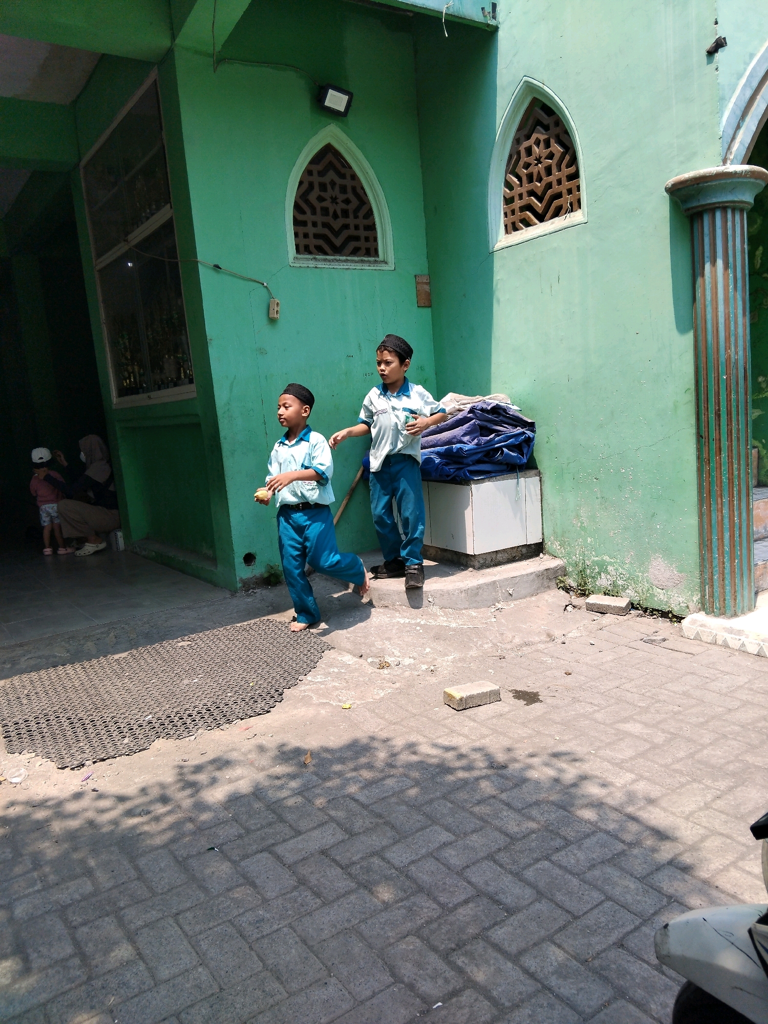 The image depicts an outdoor scene in front of a green building, likely a mosque or religious school, located in Kepuh Kiriman, Indonesia. Two young boys in light blue shirts, black caps, and blue pants are the primary subjects. The boy on the left is walking and holding a yellow object, possibly a toy. The boy on the right is also walking and appears to be looking towards the left. In the background, several people are partially visible near an entrance to the building. The building itself has distinctive arched windows with decorative latticework and a green, weathered facade. A concrete pillar with vertical blue and brown stripes is visible to the right of the frame. The ground is paved with interlocking gray bricks, and a dark mat with a circular pattern is laid out in the foreground. Shadows from trees are cast across the ground, indicating a sunny day. The scene appears to be from a public space, possibly during daylight hours.