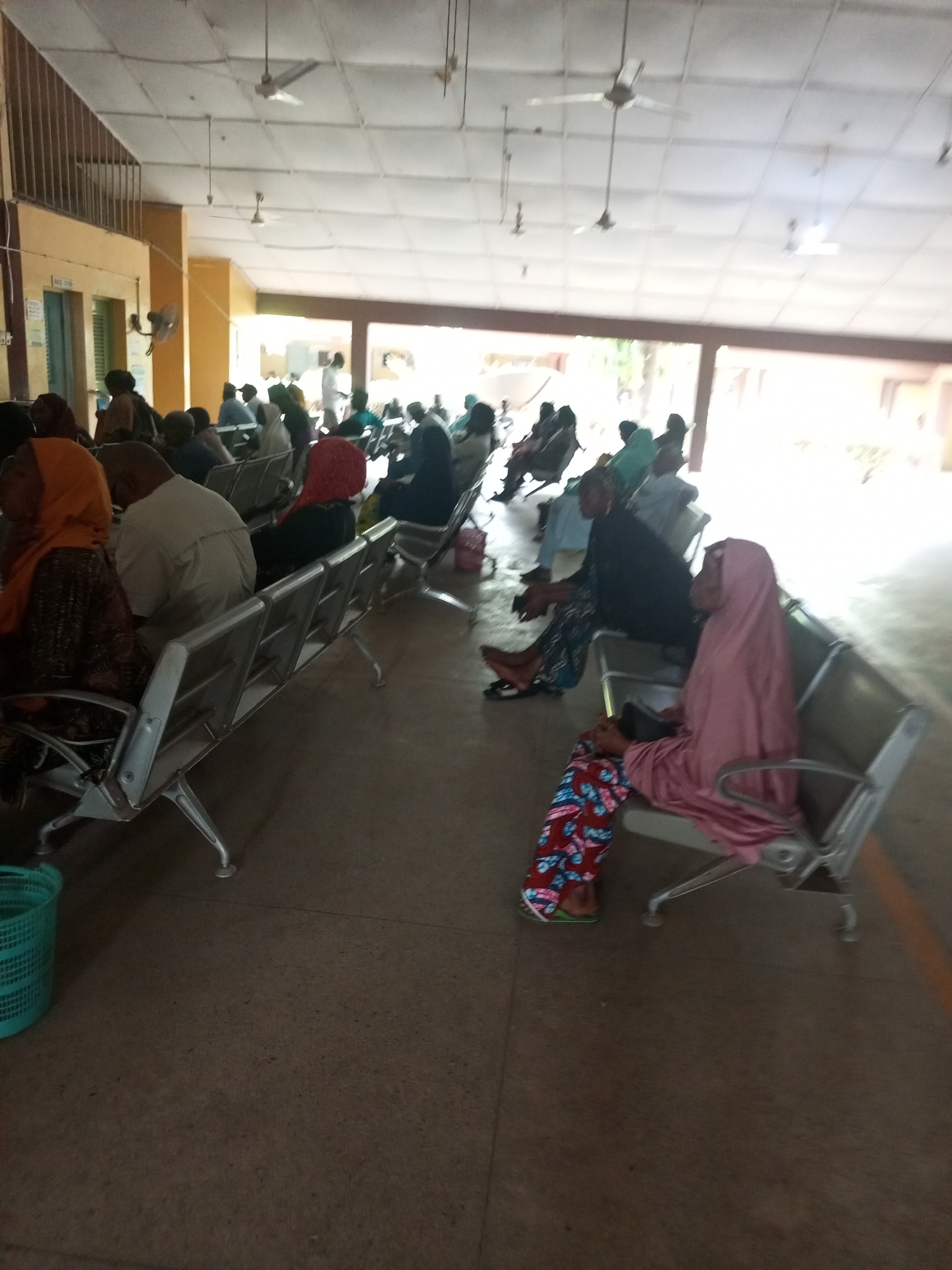 The image depicts a waiting room in Gombe, Nigeria, filled with people seated on metal benches. The room has a high ceiling with multiple ceiling fans. The environment appears to be a public building, possibly a hospital or clinic. People of various ages and genders are present, some wearing traditional Islamic clothing, like the woman in a pink hijab on the right. Light streams in from an open area at the far end of the room. There is a light green basket on the left side of the image.