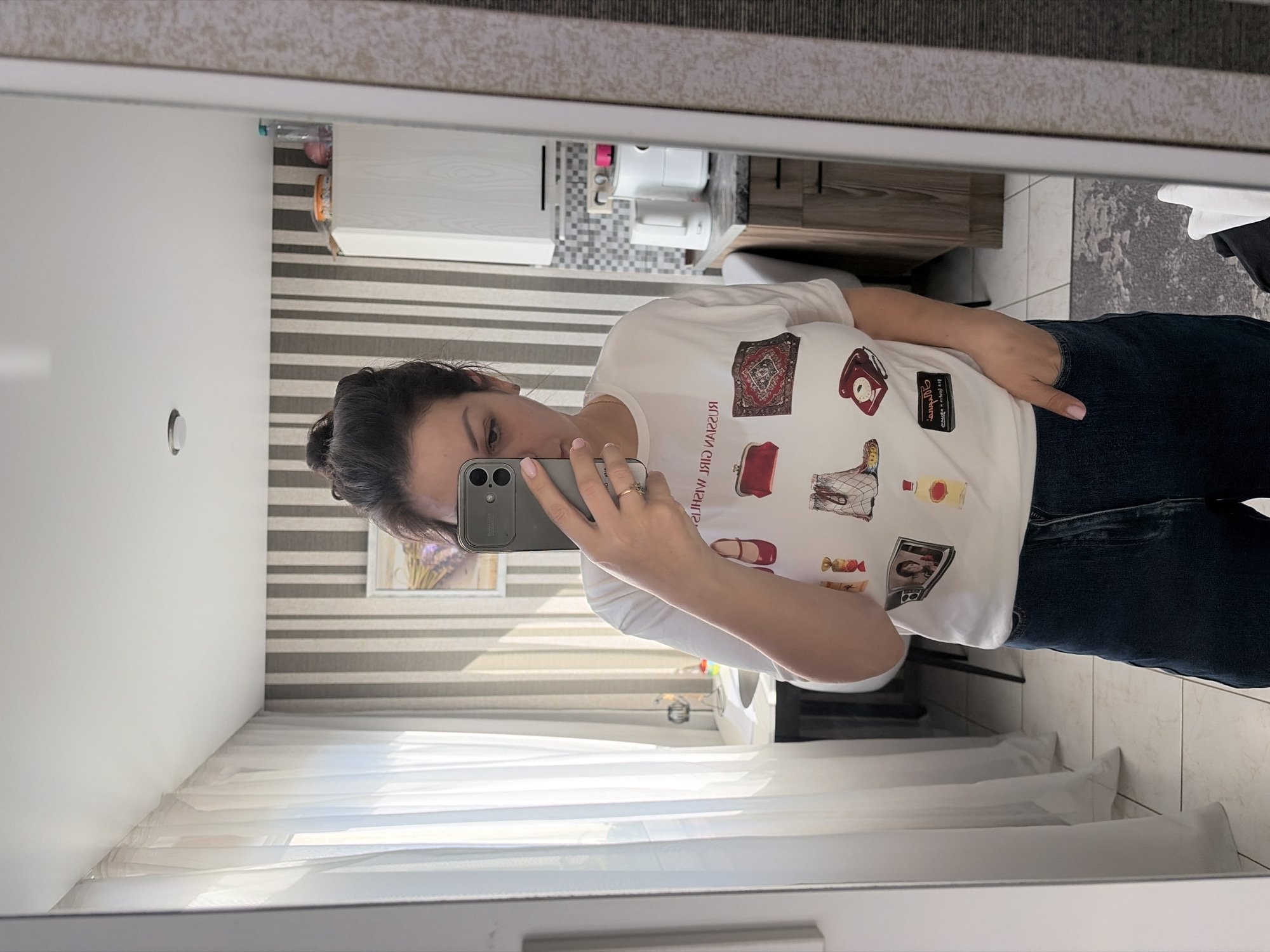 A woman is taking a selfie in a mirror. She is wearing a white t-shirt with various printed images and dark denim jeans. The t-shirt has images of a red shoe, a red purse, a rug, a boot, a phone, a bottle, a framed picture, and candy. The woman has dark hair pulled up in a bun and is wearing a ring on her left hand. Her phone is held up to take the picture.
The background appears to be an interior space, possibly a home or apartment in Sochi, Russia. The room is decorated with striped wallpaper in shades of grey and white. There is a window with white sheer curtains on the left side of the frame, letting in natural light. In the reflection, a kitchen area is visible with white cabinets, a countertop, a kettle, and other appliances. The floor is tiled.