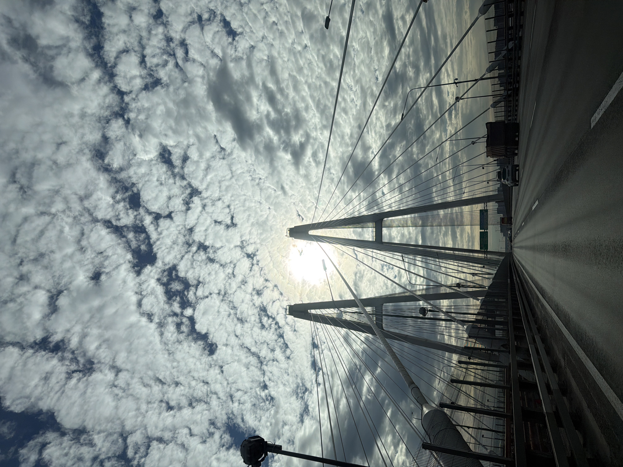 The image shows a view from a car driving on a highway bridge towards two tall, modern suspension bridge towers. The sky is filled with scattered, patchy clouds, with the sun shining brightly between the towers, creating a lens flare effect and casting shadows. The bridge deck is wet, suggesting recent rain or morning dew. Several vehicles, including a truck and cars, are visible on the bridge. The scene appears to be in an urban environment, indicated by the presence of streetlights and traffic signs in the distance. The overall atmosphere is dramatic due to the contrast between the bright sun and the darker clouds and bridge structure.