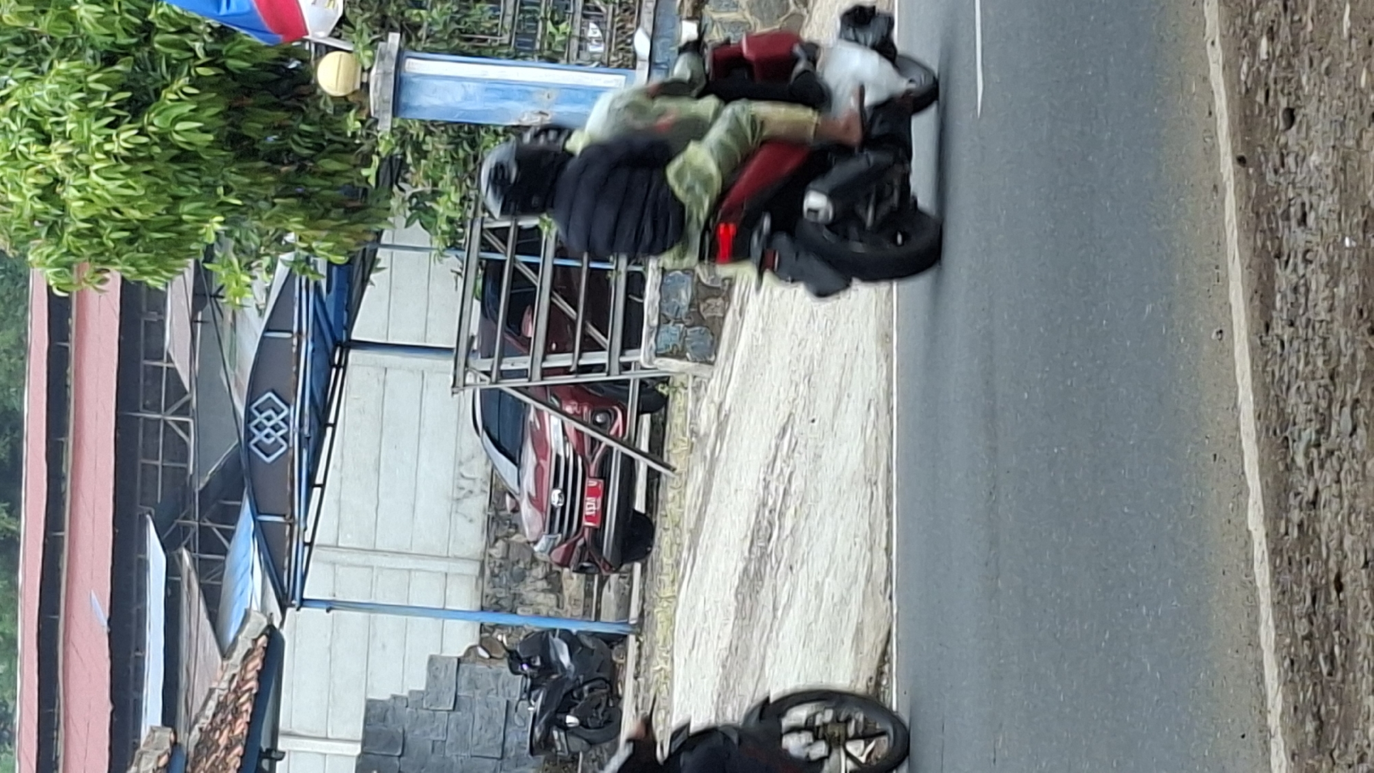 The image depicts a street scene in Cilaku, Indonesia. In the foreground, a motorcycle with two occupants is traveling on the road. The driver wears a black helmet and a dark jacket, while the passenger is wearing a light-colored garment. The motorcycle is red. Behind them, a red car is parked on a paved area, which slopes upwards. The car has a license plate that appears to read "L573 U". To the left of the car, another black motorcycle is parked. The background shows buildings with red and blue roofs, a concrete wall, and lush green trees. A blue and white pole with a yellow sphere is visible to the right, possibly part of a fence or gate. The road surface is asphalt with a white line marking the edge. The overall environment suggests a typical street in a residential or commercial area.