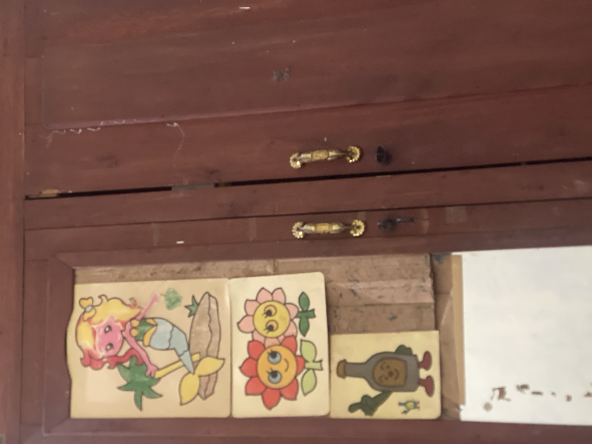 In the city of Anggaswangi, Indonesia, an aged wooden cabinet features three decorative decals affixed to its left door. The topmost decal depicts a cartoon mermaid with blonde and red hair, a green top, and a blue tail, positioned beside a palm tree and resting on a brown rock. Below the mermaid is a decal of two cartoon flowers, one red and one pink, each with wide blue eyes and smiling expressions. The bottom decal shows a cartoon bottle with arms and legs, wearing red shoes, giving a thumbs-up gesture, with a small blue and yellow creature to its left. The cabinet's right door is mostly visible, showing its dark red wooden surface and two ornate gold handles, one of which appears to have a key inserted. The overall environment suggests an indoor setting, possibly a home or shop, with the cabinet being a prominent fixture.