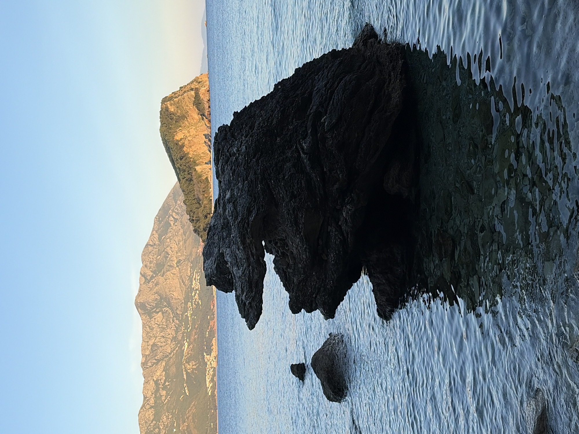 The image, when viewed upright, presents a tranquil coastal landscape near Budva, Montenegro, captured during either sunrise or sunset. In the immediate foreground, a large, dark, rugged rock formation dominates the right side, extending into the water. This rock exhibits an irregular, textured surface with distinctive overhanging sections and smaller, partially submerged rocks at its base.
The midground features calm, blue-grey sea water, reflecting the sky and light with gentle ripples across its surface. In the background, majestic mountains and cliffs rise, illuminated by a warm, golden light that suggests either early morning or late afternoon. These mountains appear to be covered in sparse vegetation, and faint glints of light on their slopes hint at distant buildings or settlements. The sky above the mountains is clear and a soft light blue.
The scene conveys a sense of natural beauty and serenity, without any specific human activity or visible text. The warm lighting contrasting with the cool tones of the water and dark rock creates a picturesque and peaceful ambiance.