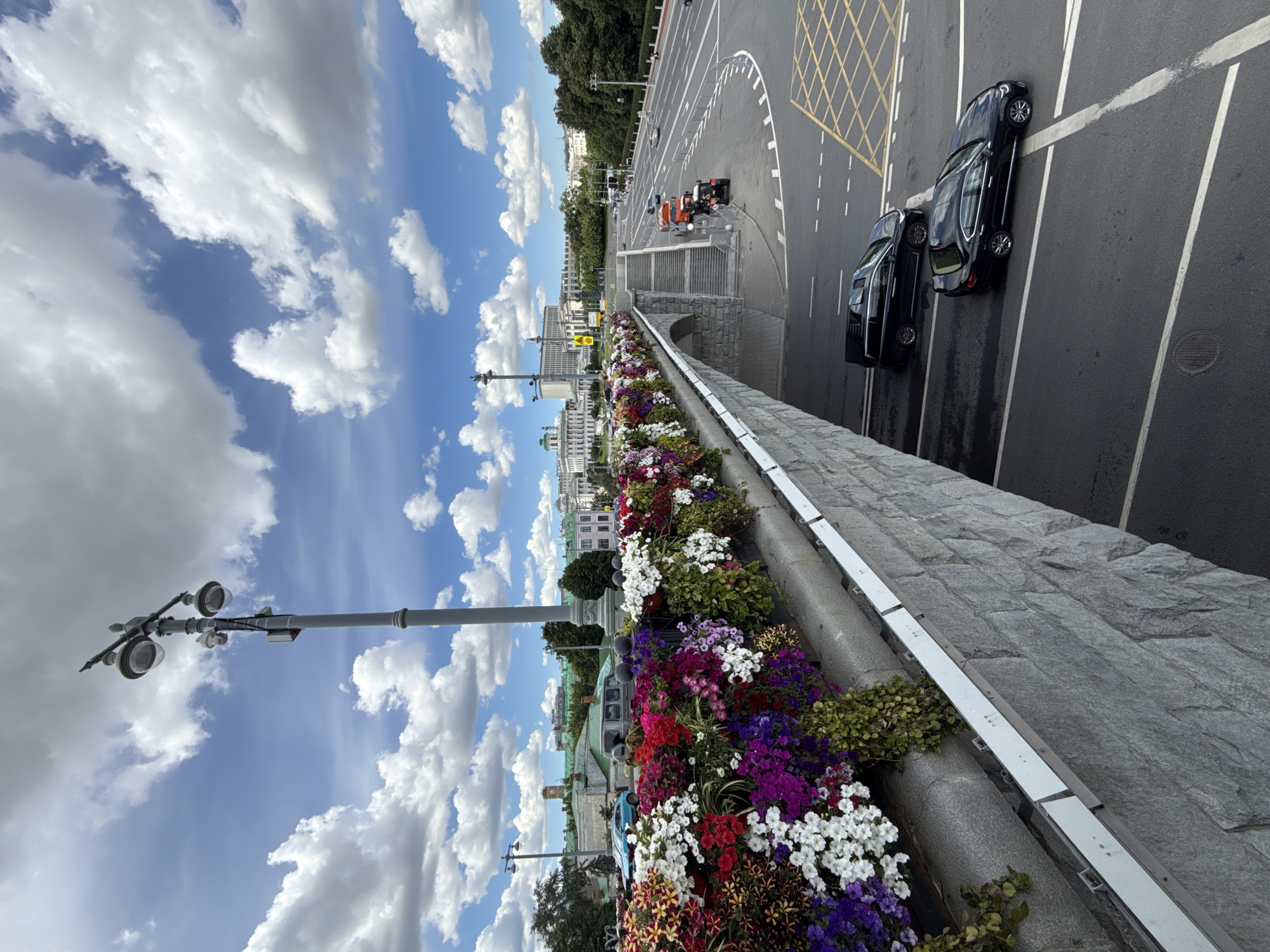 This image captures a view of the Moskvoretskaya Embankment in Moscow, Russia, on a cloudy day. In the foreground, a stone bridge is adorned with a vibrant display of colorful flowers, including red, pink, purple, and white blooms, cascading over the edge. A tall lamppost stands near the center of the bridge. A multi-lane road runs parallel to the embankment, with several cars, including a black sedan and a black SUV, navigating the traffic. In the background, several buildings, including a prominent white structure with columns and a tall tower, are visible along with trees and a clear blue sky dotted with white clouds.