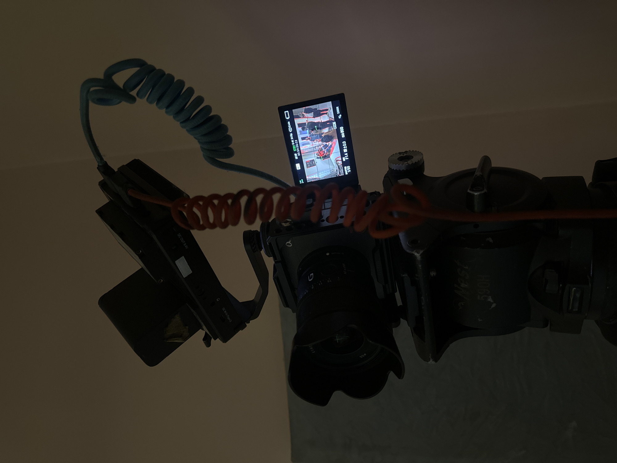 The image shows a close-up of a camera setup, likely for recording. A professional video camera, identified by the "alpha" logo and a lens with "G" and "FE 1.4/35" markings, is mounted on a Nest 590H tripod head. Attached to the camera's hot shoe is a small monitor displaying a scene with several people. A coiled orange cable connects to the monitor, and a blue coiled cable extends from the camera. The background is a plain, neutral wall, suggesting an indoor setting. The lighting is dim, casting long shadows and highlighting the equipment. There is no visible indication of the location being in Pakuro, Nigeria.