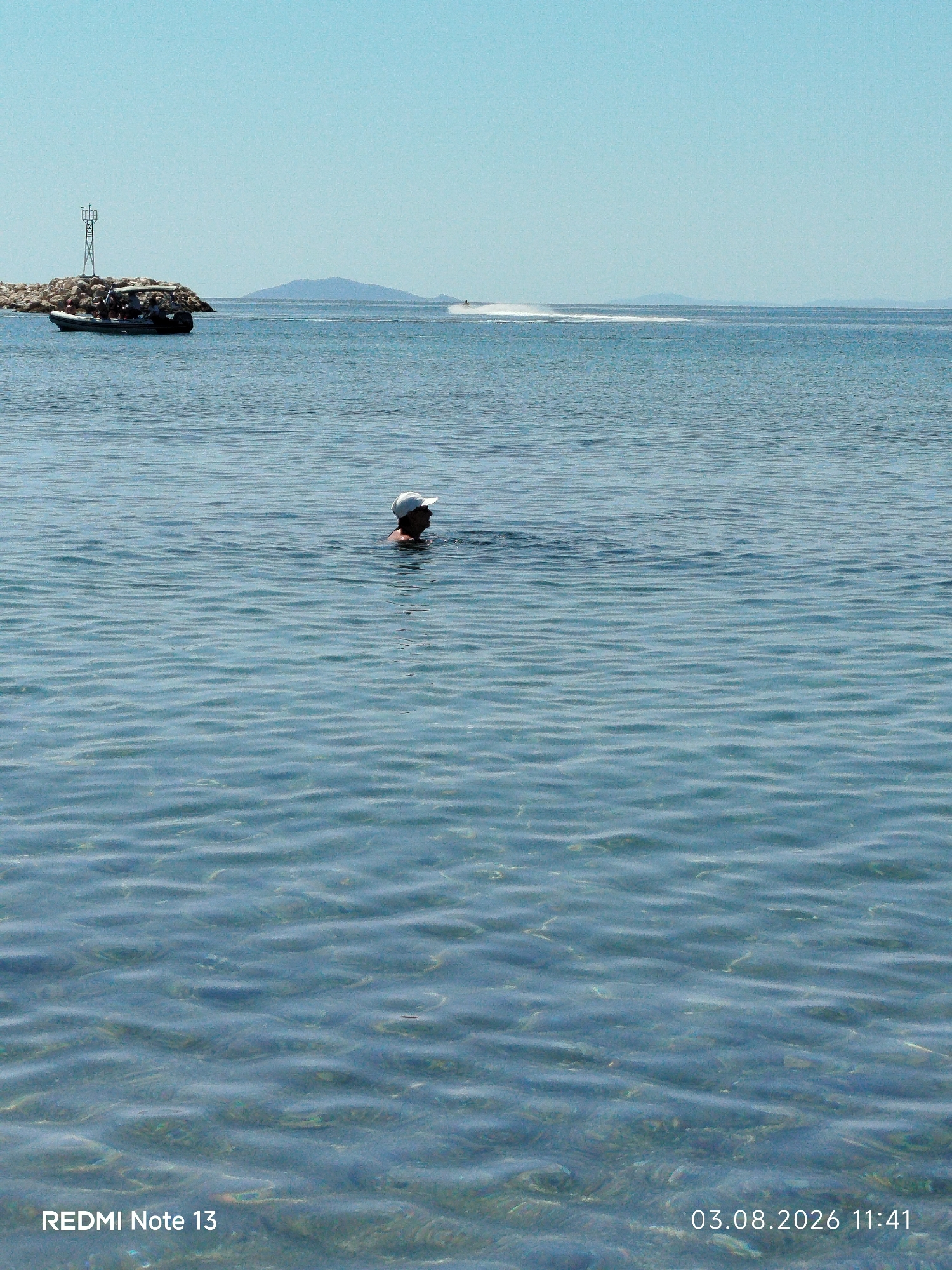 In the image, the clear, blue water of the Aegean Sea stretches towards the horizon under a cloudless sky. A person wearing a white hat is swimming in the middle ground, their head and shoulders visible above the surface. In the distance, a jet ski leaves a white wake across the water. To the left, a small boat is docked near a rocky outcrop topped with a navigational marker. The bottom right corner of the image displays the text "REDMI Note 13" and a date and time stamp: "03.08.2026 11:41". The scene appears to be a coastal area within Sithonia Municipal Unit, Greece.