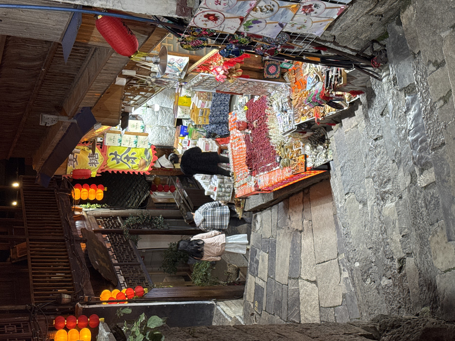The image presents a nighttime scene in the city of Furong, China, depicting a descending stone staircase leading into a marketplace. Three individuals are observed on the staircase, progressing towards the market stalls. The leftmost person wears a light-colored jacket and a white skirt. The middle person wears a plaid jacket. The rightmost person, dressed in dark attire, stands closer to the market area, facing away from the camera.

The environment is illuminated by artificial light sources, including numerous red and orange traditional Chinese lanterns hanging from the architectural structures. On the right side of the staircase, a market stall displays a large, rectangular table with an illuminated reddish-orange edge, laden with small, reddish, dried items and various packaged goods. Below this table, additional merchandise is visible. To the right, a vertical display rack holds multiple circular decorative fans with floral patterns, alongside hanging ornaments.

In the background, a shop entrance is marked by a yellow banner displaying Chinese characters for "Tea" and "Candy." This shop is brightly lit, showcasing various packaged products. The surrounding architecture consists of traditional multi-story wooden buildings featuring dark tiled roofs, ornate wooden railings, and balconies that line the narrow street. The stone staircase in the foreground is composed of irregularly shaped flagstones.