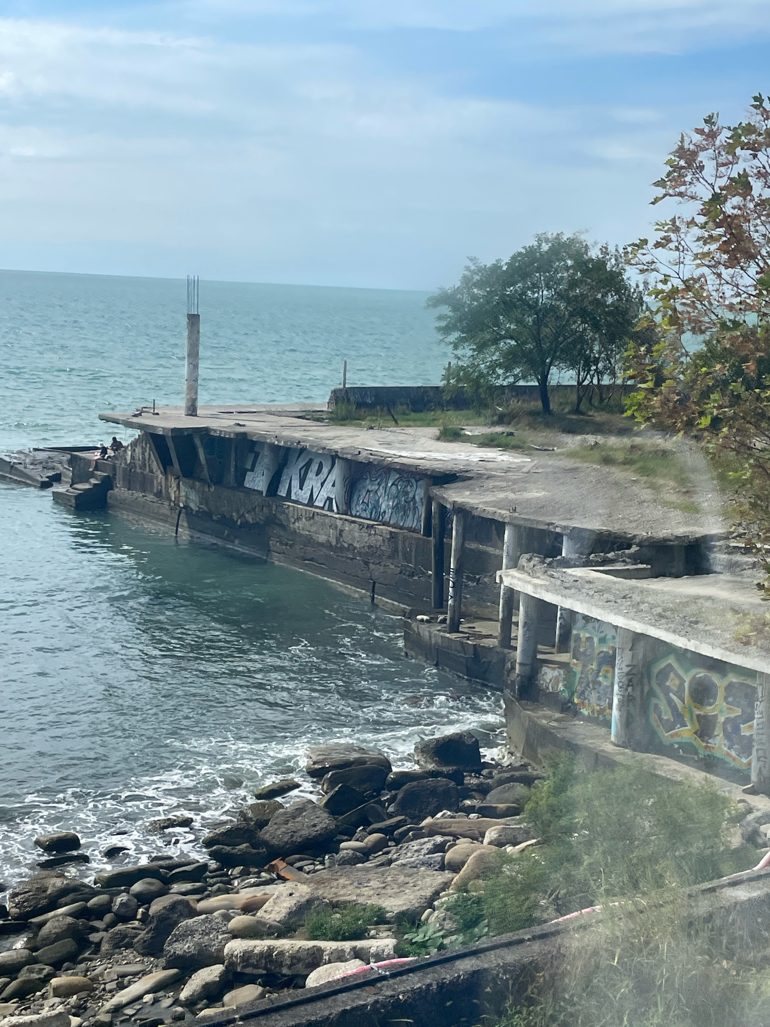 The image shows a dilapidated concrete structure extending into the Black Sea, near the coast of Sochi, Russia. The structure appears to be a pier or breakwater, with sections broken and overgrown with vegetation. Graffiti is visible on the concrete surfaces. The sea is calm, with waves gently lapping against the rocky shoreline. The sky is overcast, with a few scattered clouds. In the distance, trees and a small pole are visible. Two individuals are seated on the edge of the structure.