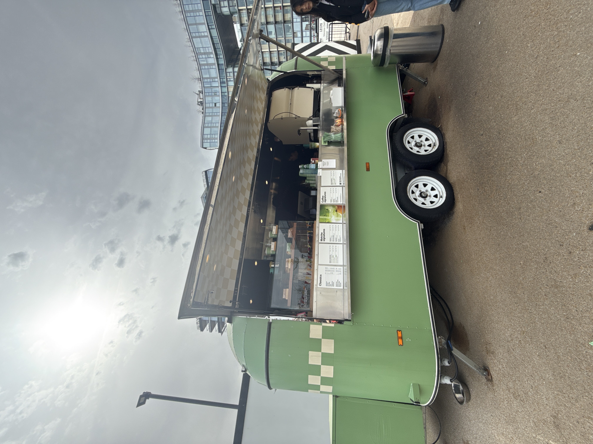 A food truck selling beverages is parked on a paved surface. The truck is primarily green with white checkered accents and has a hinged side that is open, revealing the interior preparation area and a menu board. Two tires with white rims are visible. A person stands near the front of the truck, partially out of frame. Buildings are visible in the background, suggesting an urban setting. Based on the branding "LEON" and the architectural style, this appears to be a location in the city of London, United Kingdom. The sky is overcast.