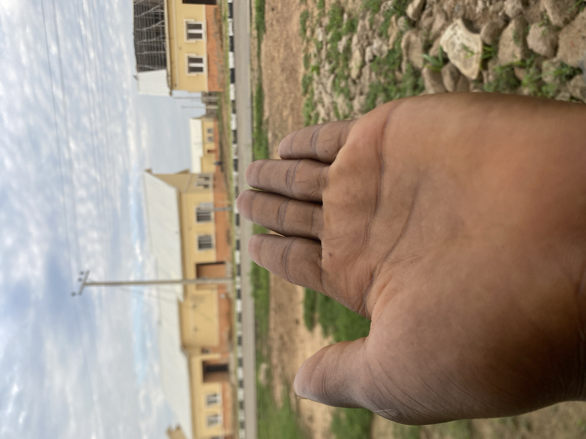 In Dumnga, Nigeria, an outdoor scene unfolds with a focus on a person's hand, palm up, in the foreground. The hand, exhibiting brown skin and visible lines, is positioned to occupy the lower left portion of the frame. Beyond the hand, the environment transitions to an unpaved area with scattered rocks and sparse greenery. Further in the background, a paved road with black and white markings runs horizontally across the image, separating the foreground from a row of yellow buildings. These structures appear to be residential or perhaps administrative buildings, featuring white roofs and numerous windows. A telephone pole stands near the buildings, and power lines stretch across the sky. The sky itself is overcast with a textured pattern of clouds. The overall impression is one of a developing area, with construction or recently completed structures in an outdoor, possibly semi-arid, setting.