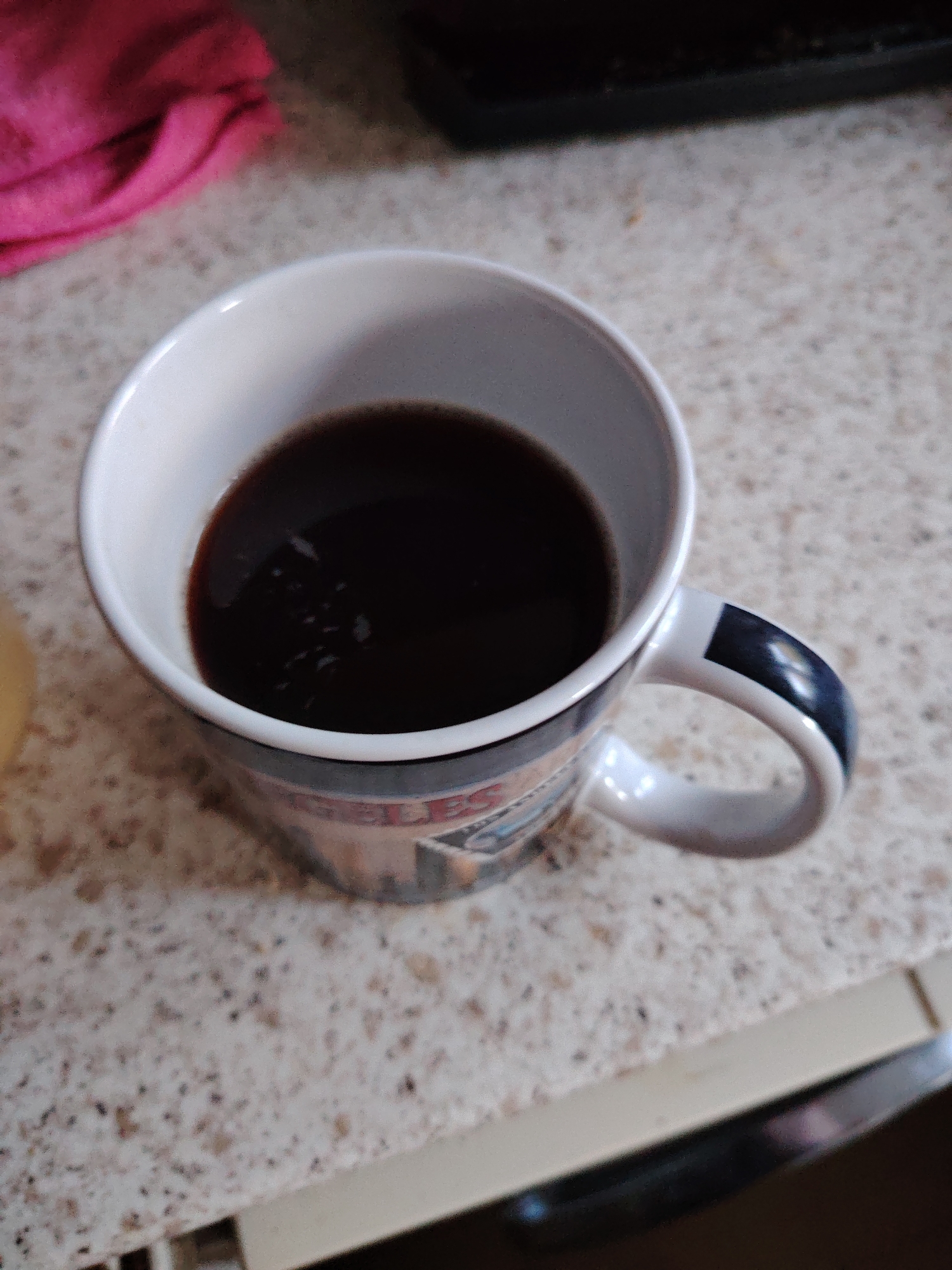 The image shows a close-up, high-angle shot of a white mug filled with dark liquid, likely coffee, resting on a speckled countertop. The mug has a blue and white design on its exterior and a blue stripe on its handle. To the upper left of the mug, a pink cloth is visible. The countertop has a speckled pattern of light and dark specks. The background is out of focus, but appears to be a kitchen or dining area. There are no people visible in the image. The scene is static and there is no indication of any action taking place. The lighting is adequate and appears to be from an overhead source. The image quality is good, with clear focus on the mug and its contents. The location is not specified in the image or its context.