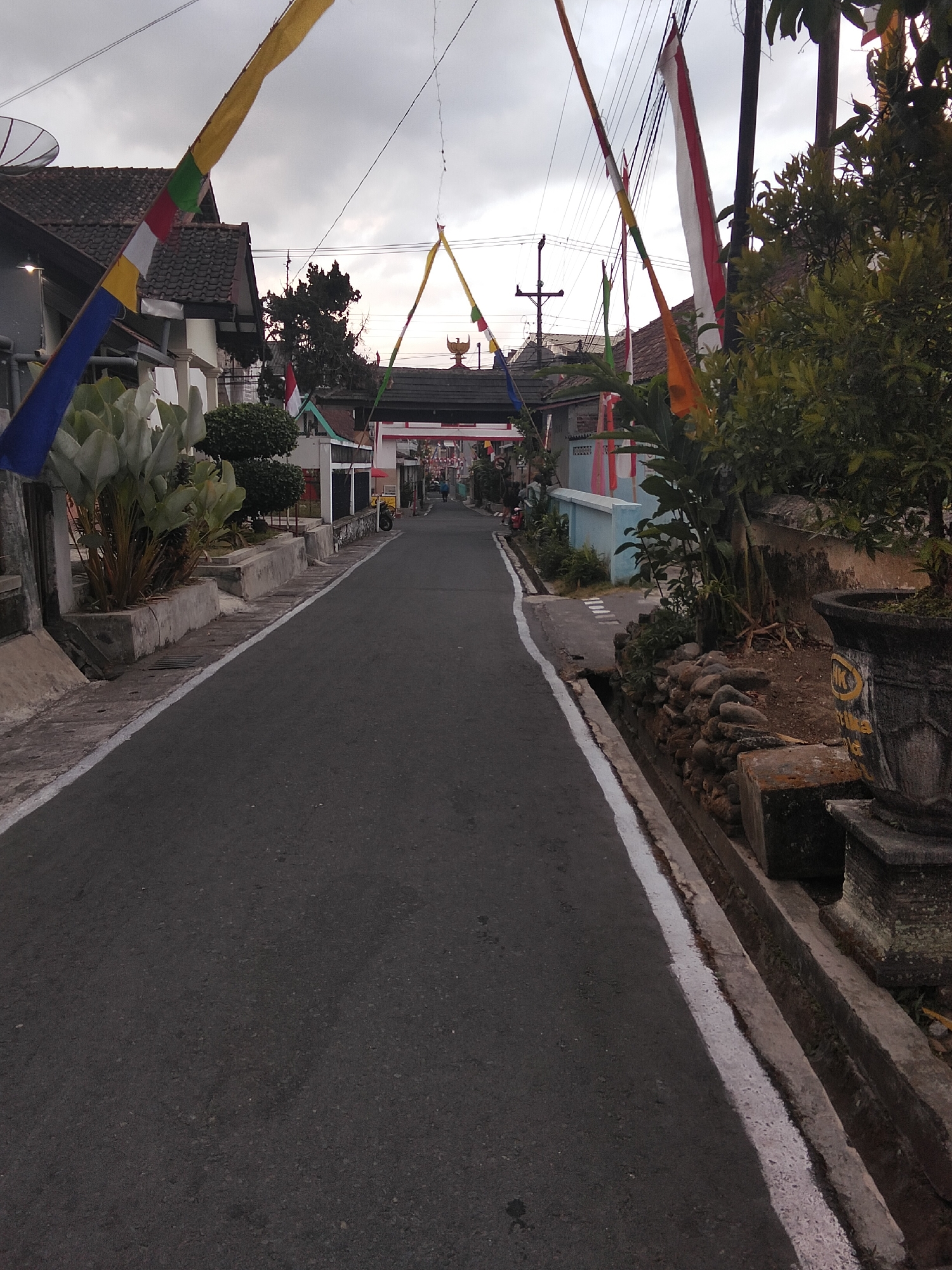 The image depicts a street scene in Magelang, Indonesia, likely on a cloudy day. A paved road runs through the center, bordered by a concrete drainage ditch with a white line along the edge. On the left side of the road, houses with tiled roofs and decorative flags are visible. One house features a satellite dish. Plants, including a large-leafed one in a planter, are also present. On the right side, a stone-lined embankment leads to a tree and a large decorative pot. In the background, a red and white archway or gate spans the road, adorned with more flags. A few people are visible in the distance, walking and on a motorcycle, suggesting a residential or community area. The overall impression is that of a quiet street decorated for a local event or celebration.