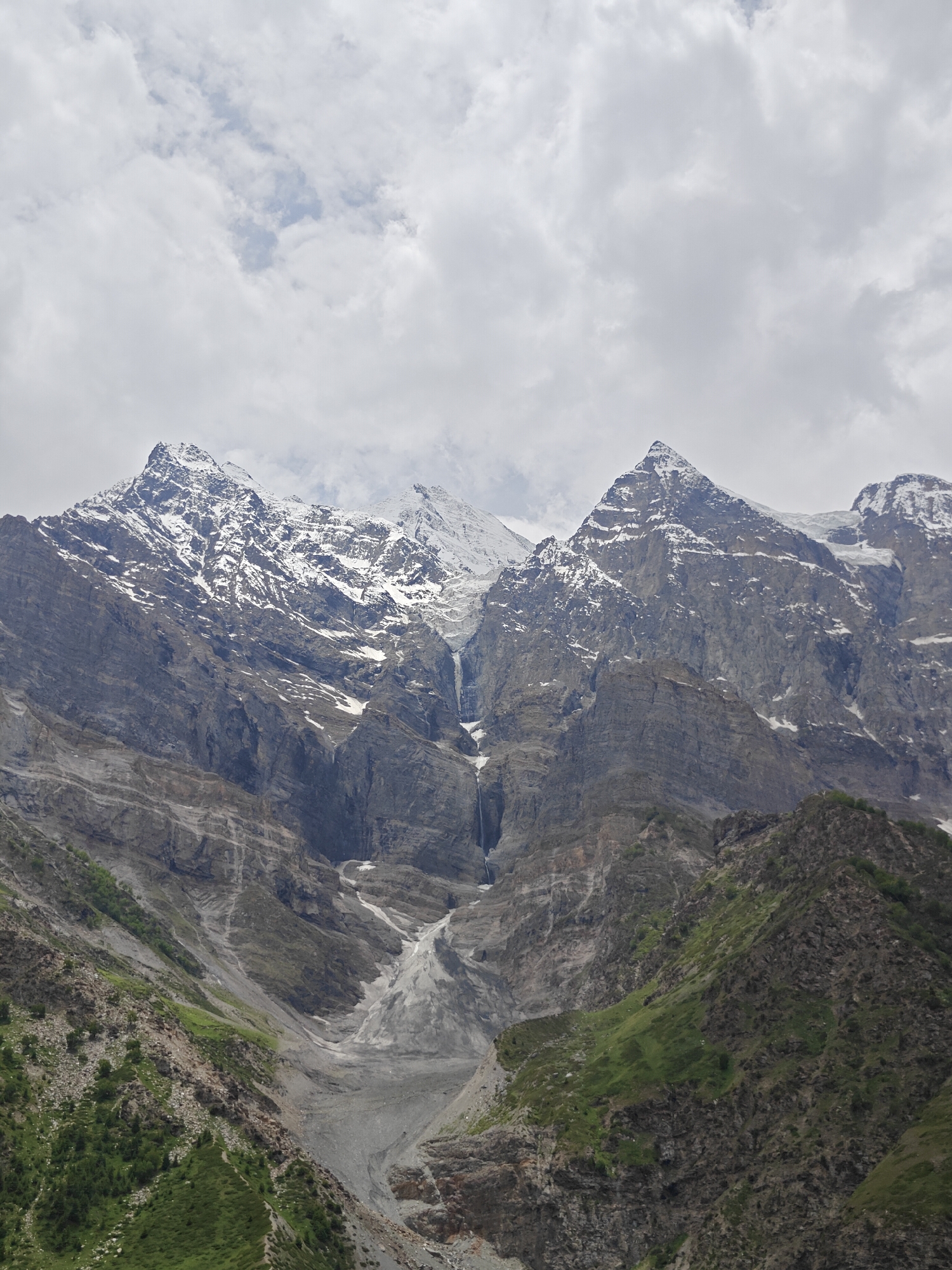 The image depicts a majestic mountain range with snow-capped peaks and rugged slopes. A waterfall cascades down the central mountainside, feeding into a glacial valley. The lower slopes are covered in green vegetation and rocky outcrops. The sky above is filled with dramatic, cloudy formations. The scene is likely located in a high-altitude mountainous region such as Jagle, India, known for its natural beauty and challenging terrain. There are no people or man-made objects visible in this natural landscape.
