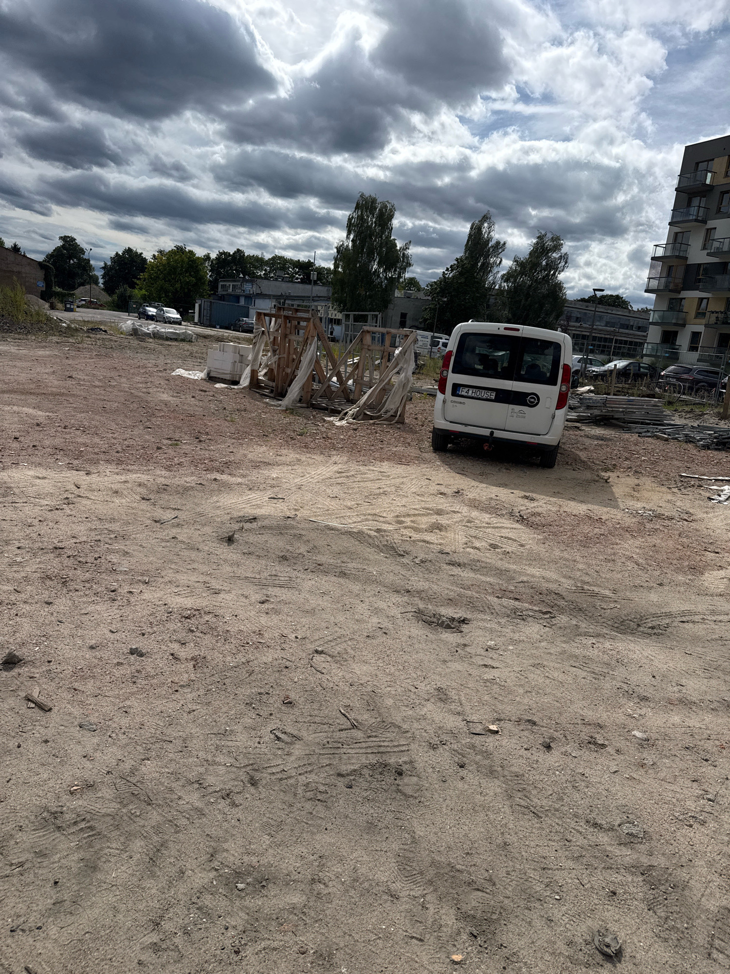 The image depicts an outdoor scene at a construction site in Nowa Sól, Poland. The foreground is dominated by a dusty, uneven ground surface with visible tire tracks and scattered debris. In the mid-ground, there is a white van with the license plate "F4 HOUSE." Next to the van are piles of construction materials, including wooden pallets, plastic sheeting, and stacks of white blocks. Further in the background, several cars are parked. The environment includes trees and buildings, with a large, modern residential building on the right side of the frame. The sky is overcast with dramatic clouds.