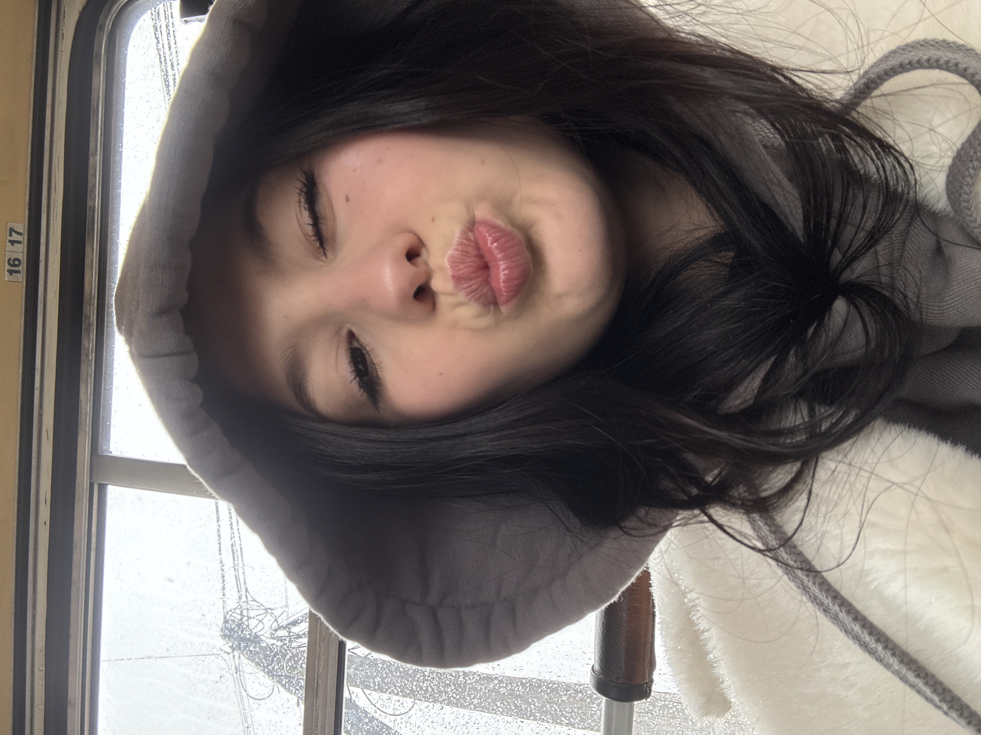 A young woman with dark hair is posing for a selfie near a window on what appears to be a rainy day. She is wearing a gray hooded sweatshirt and a white, fuzzy jacket. The woman is pouting her lips towards the camera and her eyes are partially closed. A brown object is partially visible below the hood. A number 16 and 17 are on the window frame. It is not possible to verify the location in Фоки, Russia from this image.