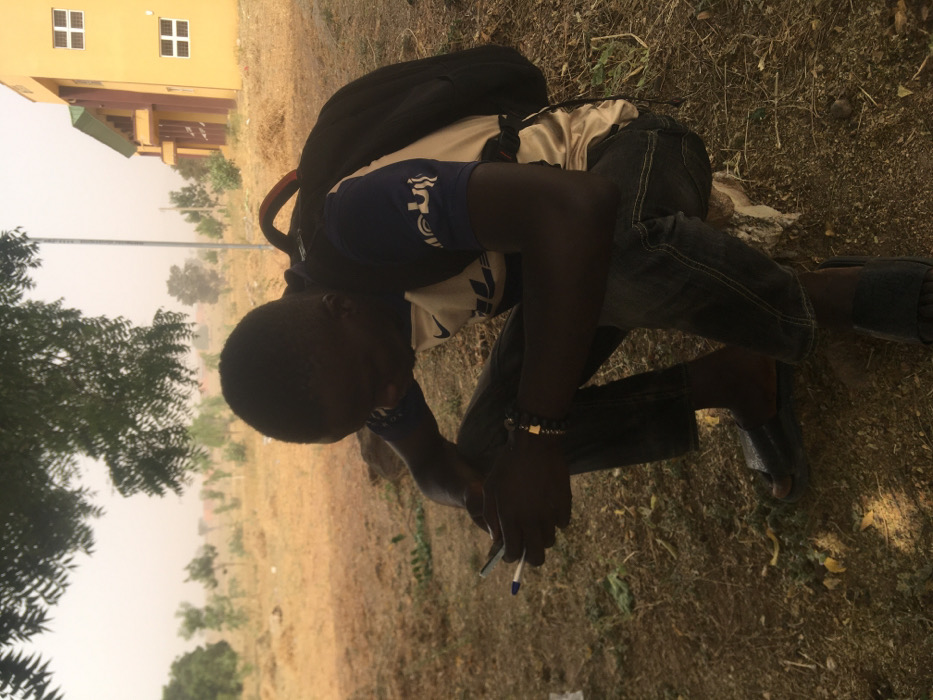 The scene in Pindiga, Nigeria, depicts a dark-complexioned male squatting on dry, light brown soil under the partial shade of a tree. He is wearing a white short-sleeved t-shirt with navy blue sleeves and a white Nike logo, dark denim trousers, and dark slide sandals. A black backpack with orange accents is worn on his back, and a dark beaded bracelet with a light-colored accent is on his left wrist. He holds a white mobile device in his left hand and a blue pen in his right hand, his gaze directed downwards towards his hands. The immediate ground contains sparse dry vegetation and a small, light-colored rock near his right leg. The background shows a hazy, light-colored sky over an expansive, dry field with scattered small trees and shrubs. A tall, slender pole stands in the mid-distance. To the right, a multi-story building with yellow-brown walls, a green roof trim, and several white-framed windows is visible.