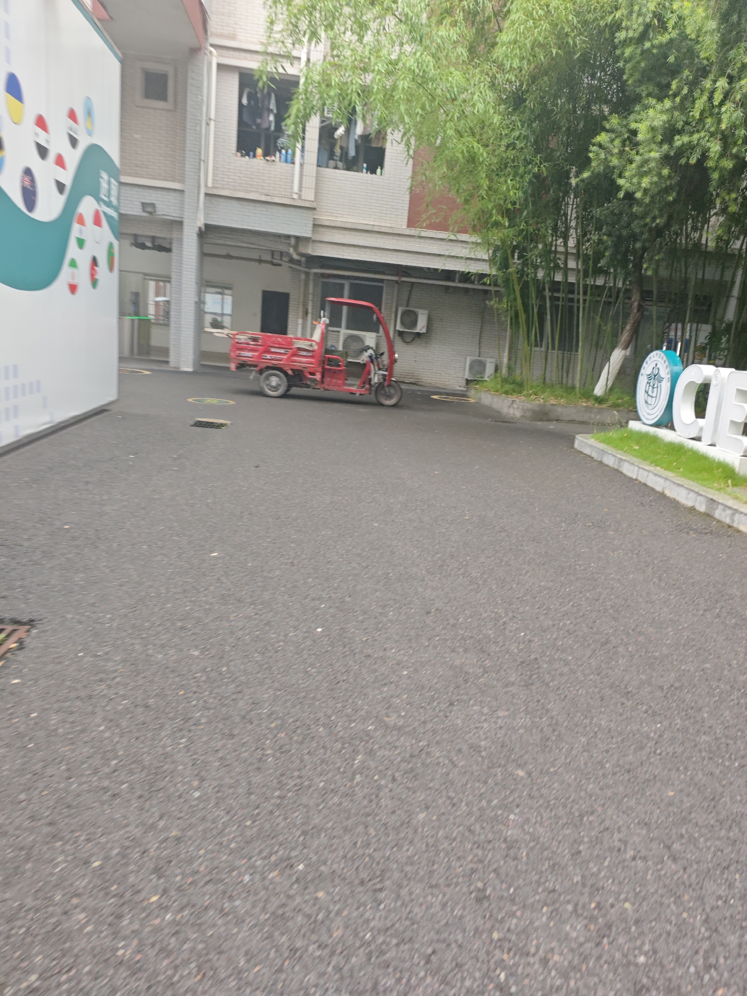 The image depicts an outdoor scene in the Chongqing High-tech Zone, China. In the foreground, a dark asphalt surface stretches across the frame, with some scattered debris and a drain grate visible on the left. In the mid-ground, a bright red three-wheeled cargo vehicle is parked on the asphalt. Behind the vehicle, a building with light-colored brickwork and windows can be seen. Green bamboo plants grow in front of the building, and further to the right, a large white sign with blue lettering and a circular logo is partially visible, suggesting it might be part of an establishment or institution. The environment appears to be an urban or semi-urban setting, likely within a business park or campus.