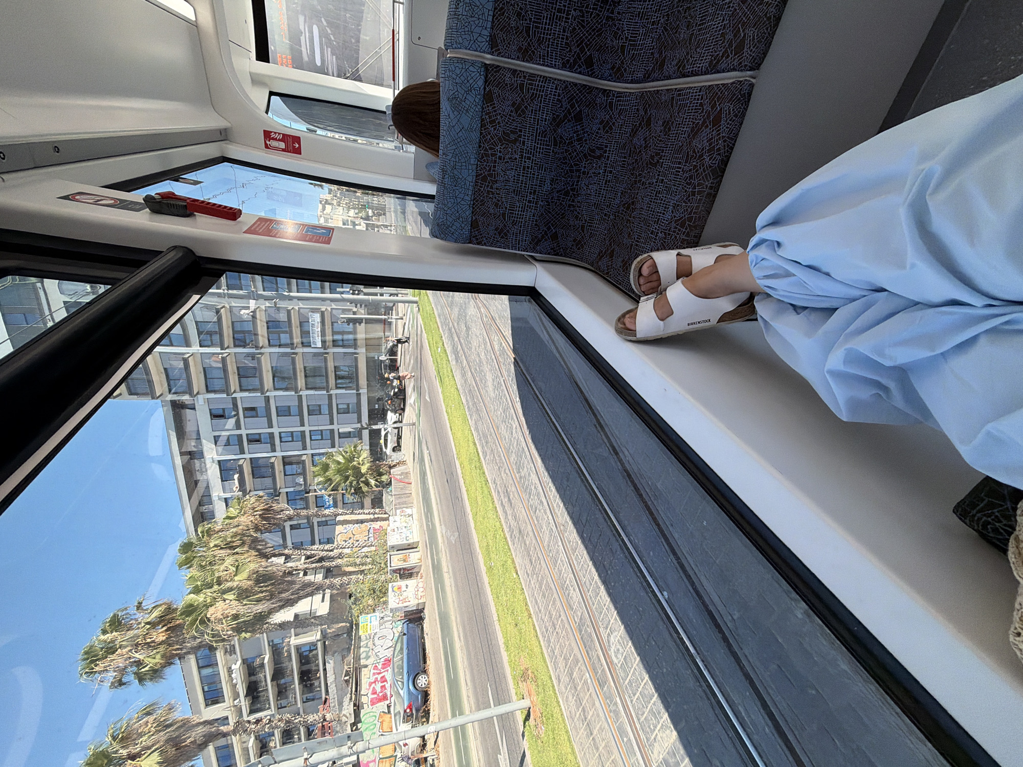 This media file shows a person's feet wearing white Birkenstock sandals, resting on the edge of a tram or train seat. The person is covered by a light blue garment, possibly a dress or robe. The scene appears to be inside a moving vehicle, as suggested by the reflection of buildings, palm trees, and tram tracks in the window. The outside environment indicates a sunny day, with a clear blue sky. The buildings outside have a Mediterranean architectural style, and the presence of graffiti suggests an urban setting in a warm climate. The tram tracks are laid out on a paved road with a strip of green grass between them.
Visible text includes "BIRKENSTOCK" on the sandals. There are also some red and white warning stickers on the interior of the vehicle, one with an icon of a person with a disabled sign, another with an arrow and a siren-like symbol. A red and grey object, possibly a tool or safety equipment, is placed on a ledge near the window.
The overall impression is one of travel and relaxation on a bright day, possibly in Tel Aviv, Israel, given the architectural style and vegetation. The composition, with the focus on the feet and the view outside, evokes a sense of personal journey and observation.