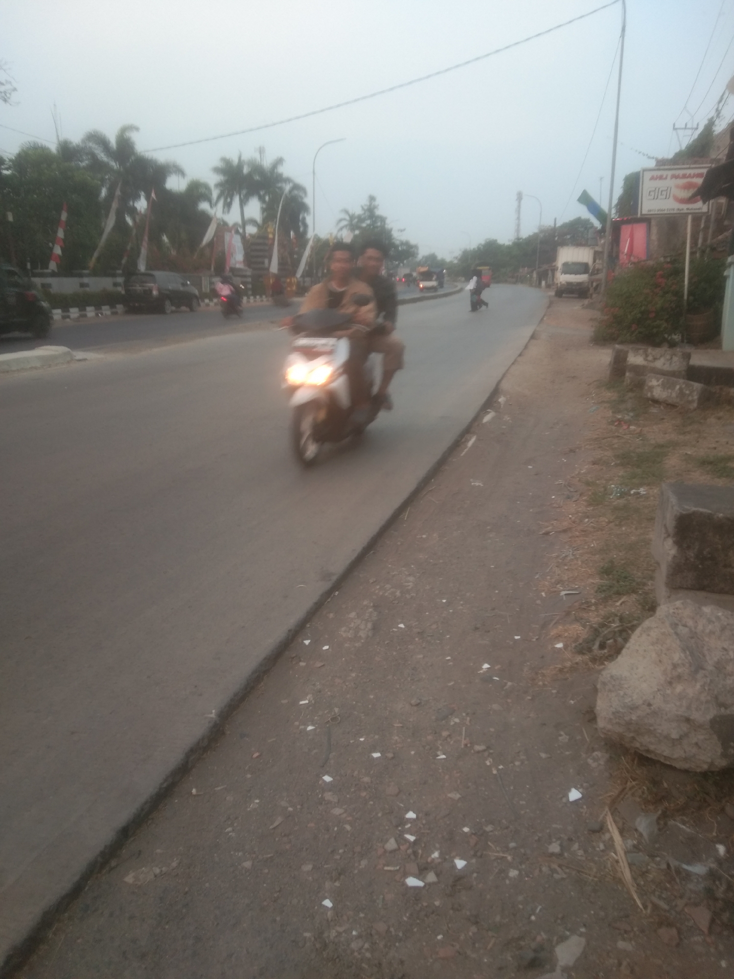 The image depicts a hazy street scene in Subang, Indonesia, likely in the early morning or late afternoon due to the diffused light. The foreground shows a paved road with a dirt shoulder. A motorcycle with two occupants is the central subject, moving away from the viewer towards the left of the frame. The motorcycle's headlights are illuminated. On the right side of the road, there is a sidewalk area with some large rocks and sparse vegetation. In the background, several vehicles, including cars and a truck, are visible on the road. Trees, some with palm fronds, line the left side of the street, interspersed with what appear to be flags or banners. Buildings and signs are visible further down the road on the right, one sign displaying text in Indonesian, possibly indicating a business. The overall atmosphere is somewhat blurry, suggesting movement or low light conditions.
