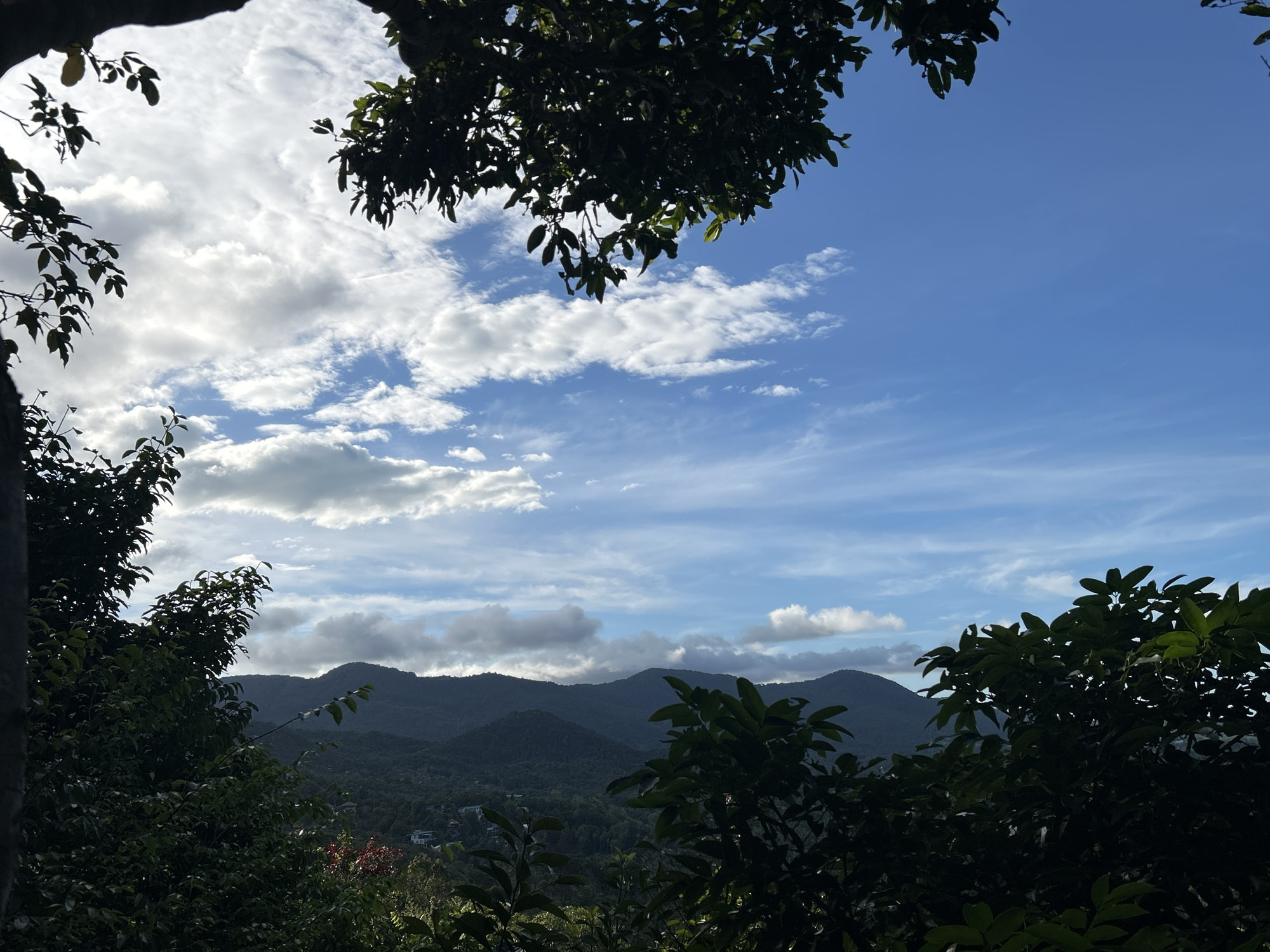 The image presents a landscape view from ตำบลเกาะพะงัน, Thailand. In the foreground, lush green foliage obscures portions of the scene, framing a mountainous backdrop. Distant mountain ranges are visible, their silhouettes against a cloudy, blue sky. The sky features a mix of cumulus and cirrus clouds, suggesting a partly cloudy day. A few buildings are visible amidst the vegetation in the mid-ground.