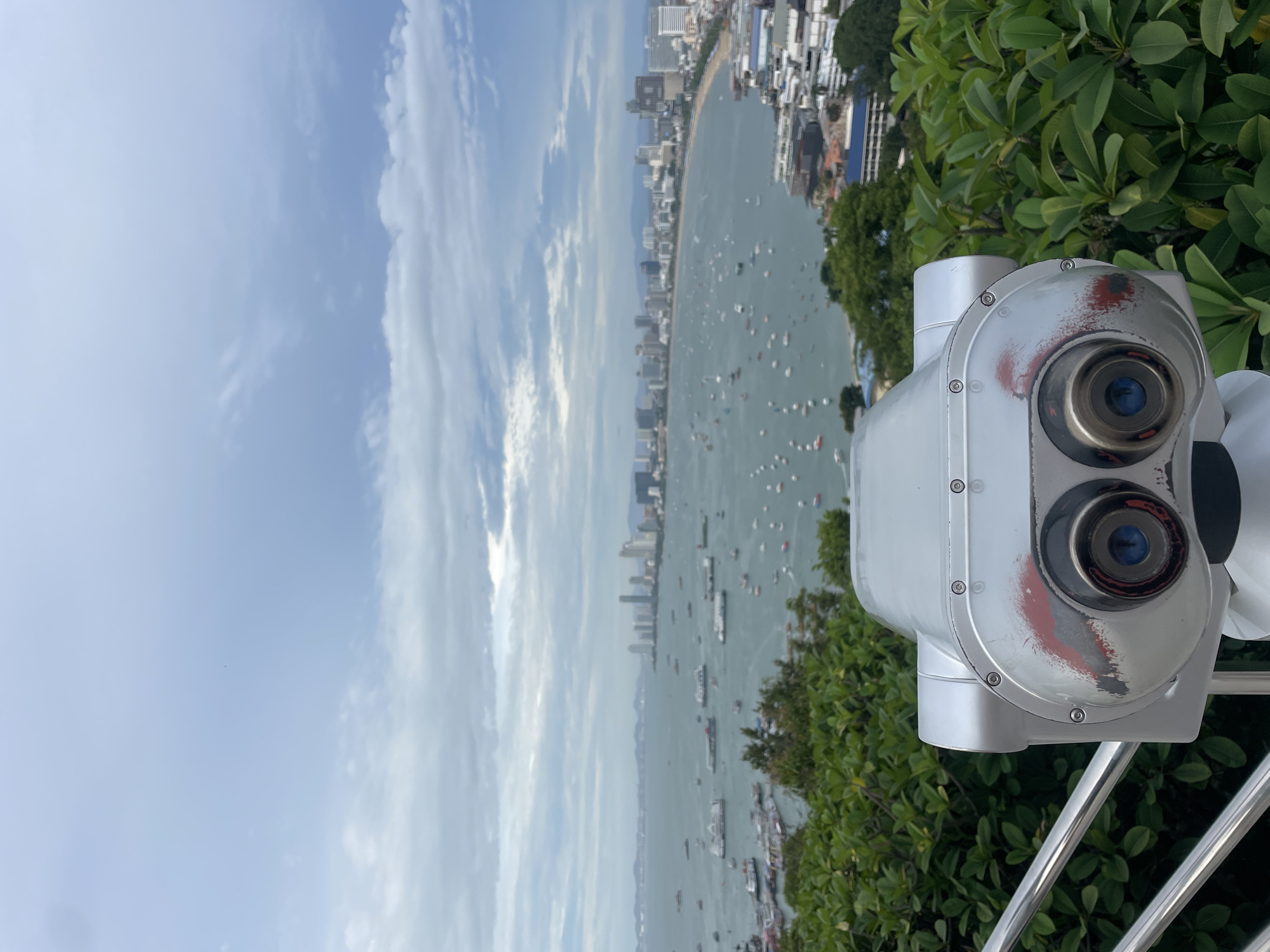 The image shows a wide-angle view from a high vantage point overlooking Pattaya Bay in Pattaya City, Thailand. In the foreground, a silver, heavy-duty tourist telescope stands on a tripod, its two lenses facing the viewer. The telescope has visible signs of wear, with red paint marks on its surface. Lush green foliage surrounds the telescope, and a silver railing is visible to its left.
The middle ground features a vast expanse of the sea, dotted with numerous boats of various sizes, including ferries, smaller vessels, and pleasure crafts. The water is calm, reflecting the overcast sky.
In the background, the cityscape of Pattaya unfolds along the coastline. A dense collection of high-rise buildings, hotels, and apartments forms the urban skyline. The city stretches along the bay, with some areas appearing more developed than others. The horizon is marked by distant hills or mountains, shrouded in a light haze. The sky is cloudy, with patches of lighter and darker gray, suggesting an overcast day with diffused light. The overall scene captures a panoramic view of the city's waterfront and its bustling bay.
