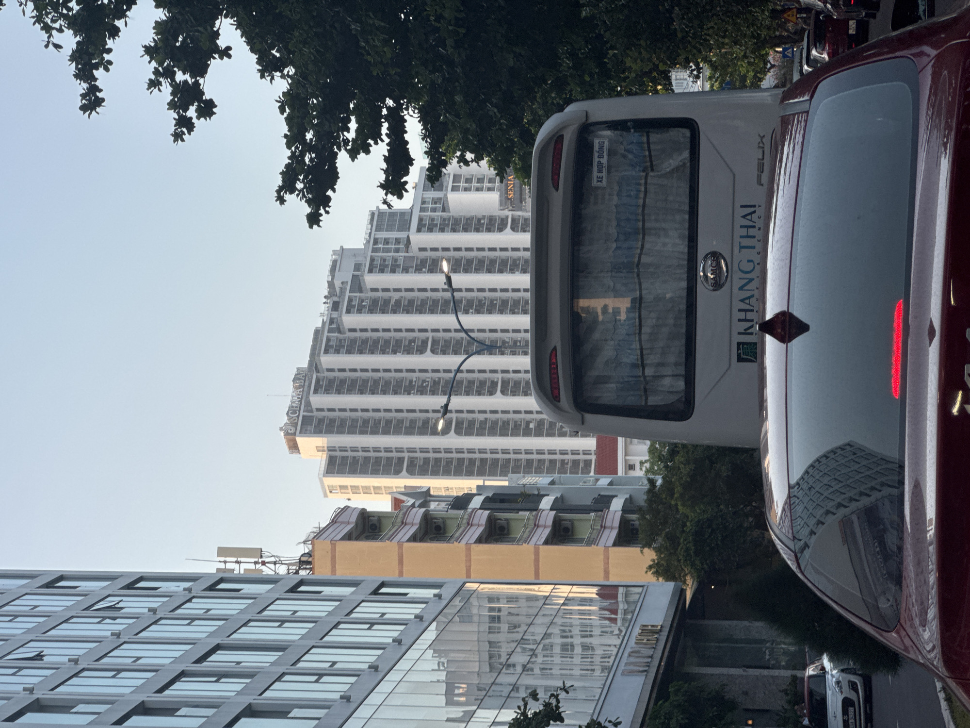 The image presents an urban street scene in Bac Nha Trang Ward, Vietnam, under clear daylight conditions, viewed from behind a vehicle.
In the immediate foreground, the rear of a dark red sedan is visible, with its central high-mounted stop lamp illuminated. A black shark-fin antenna is affixed to the trunk. The rear window reflects a portion of a grid-patterned building.
Directly in front of the sedan is the rear of a white passenger bus. The bus is branded with "SAMCO" and "KHANG THAI TRAVEL AGENCY FELIX," including an associated logo. A sign affixed above the rear window displays "XE HỢP ĐỒNG" in blue text on a white background. The bus's rear window is obscured by light blue interior curtains.
To the left of the bus, a multi-story building with a grey structural frame and extensive glass windows rises. Its facade exhibits reflections of adjacent structures and the sky. A partial sign near its base reads "NANA HOTEL." Adjacent to this glass building, an older, narrower multi-story building with a yellow primary facade, pink balcony railings, and multiple external air conditioning units is visible. Further down the street on the left, a white Suzuki Swift is parked.
The background is dominated by a cluster of tall, white, multi-story residential or mixed-use high-rise buildings. These structures feature prominent vertical architectural elements. The illuminated signage "SCENIA BAY" is visible on the rooftop of one building, and "SENIA" appears on another building further back. A street light with two lamps, one of which is illuminated, is positioned on a pole above the bus.
To the right side of the frame, a large tree with dense, dark green foliage partially obstructs the view of the background buildings and the pale blue sky. A communications antenna tower is visible on the rooftop of a building to the far left.