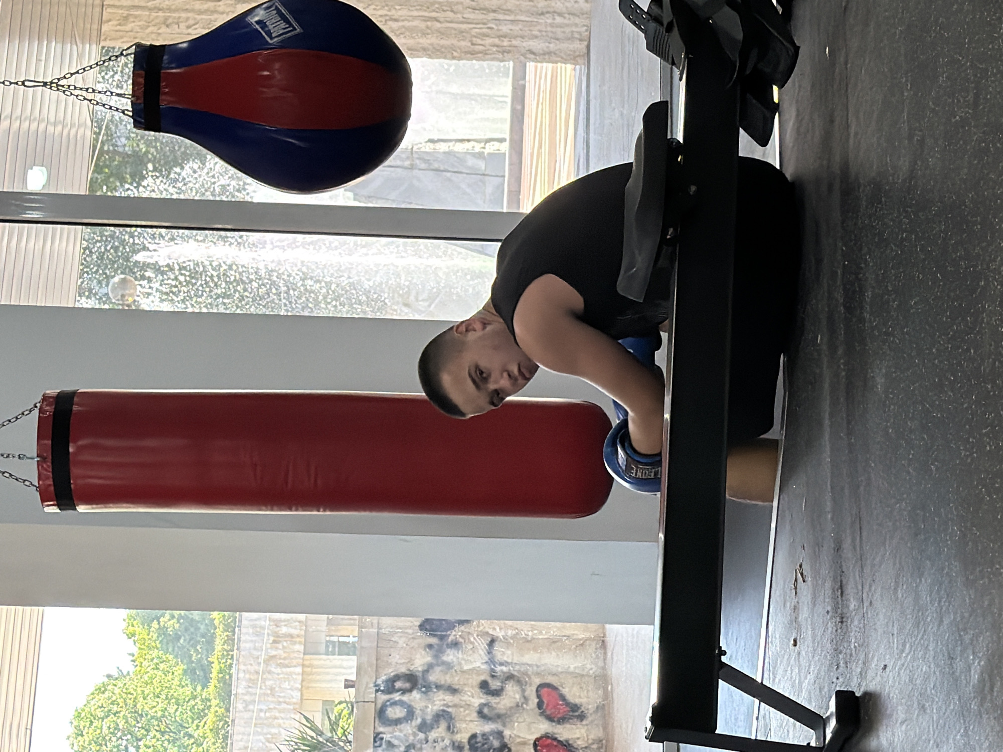 A man wearing boxing gloves stands in a gym in Blagoevgrad, Bulgaria. A punching bag and speed bag are visible in the background. He is positioned near a rowing machine.