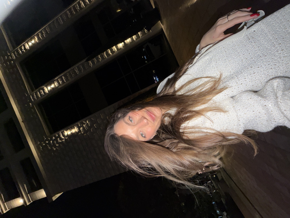 A fair-skinned woman with long brown hair and blue eyes gazes at the camera. She is wearing a white sweater with shiny accents. Her left hand, adorned with a few rings and red nail polish, is visible near her shoulder. The background is dark, featuring the exterior of a building illuminated with recessed lights above windows. The scene has low lighting and the image is rotated counterclockwise.
