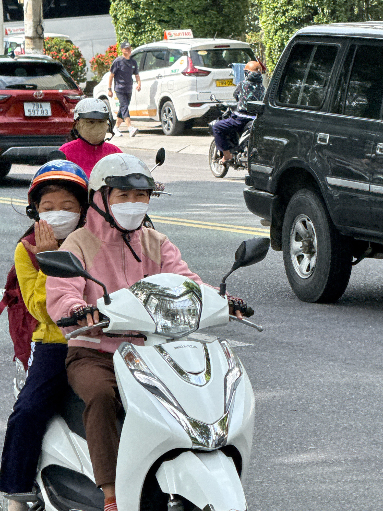This is a street-level, eye-level, medium shot captured in Nha Trang, Vietnam, showing a busy road with multiple vehicles and pedestrians. In the foreground, two individuals, presumably a mother and child, are riding a white scooter. The adult in the front wears a pink hoodie and a white helmet, with a white face mask covering their lower face. The child behind them wears a yellow jacket, a blue and orange helmet, and a white face mask. The child's right hand is held in a prayer-like gesture near their face. They are traveling on a paved road with yellow lane markings visible.
In the middle ground, a red SUV with a license plate reading "79A 597.83" is parked on the left. Behind the SUV, a white taxi with "SUNTAXI" branding is visible. Further back, a person in a dark blue uniform and cap is walking on the sidewalk, and another individual on a motorbike, wearing a patterned jacket and helmet, is also present.
The background is filled with lush green trees and foliage, indicating a sunny day. A large black SUV is positioned prominently on the right side of the frame, partially obscuring the other vehicles. The overall scene depicts typical urban traffic and pedestrian activity in Vietnam.