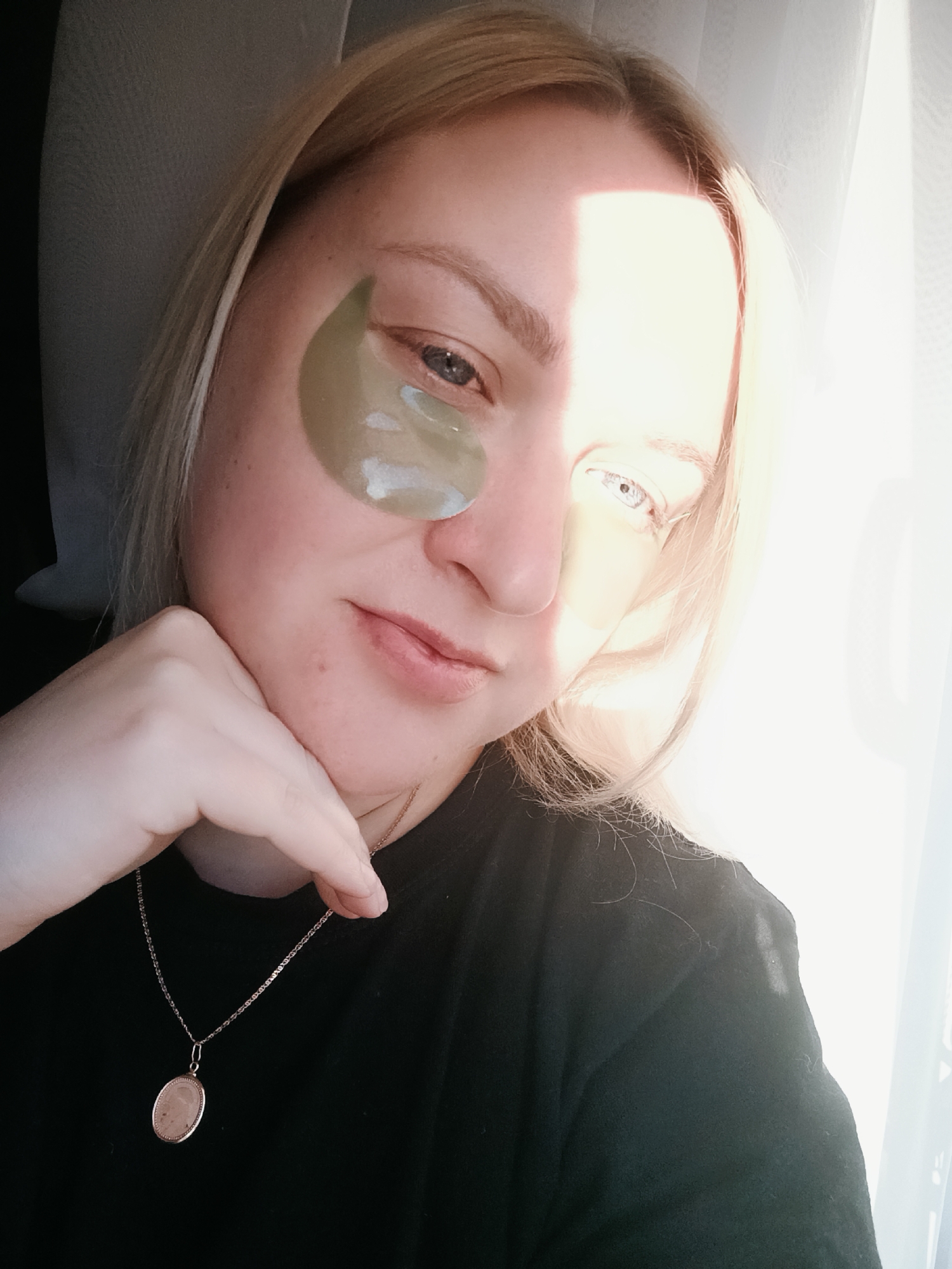 A young adult woman with blonde hair is captured in a close-up, chest-up selfie, illuminated by strong natural light from a window to her right. Her face is partially in shadow and partially brightly lit, creating a dynamic contrast. She has greenish-gold hydrogel under-eye patches applied beneath both eyes, suggesting a self-care routine. Her left eye, visible through the light, appears light in color. She has a neutral to slightly contemplative expression with a hint of a soft smile, and her gaze is directed towards the camera.
Her chin is gently resting on her right hand, which is visible in the lower left of the frame. She is wearing a plain dark top, likely a t-shirt, and a delicate gold-toned chain necklace with a small, oval-shaped pendant. Behind her head on the left, a soft, dark grey surface is visible, possibly a pillow or upholstery. To her right, a bright, translucent white curtain diffuses the sunlight, indicating an indoor setting next to a window. The overall ambiance suggests a relaxed moment, possibly in a home environment. There is no visible text in the image.