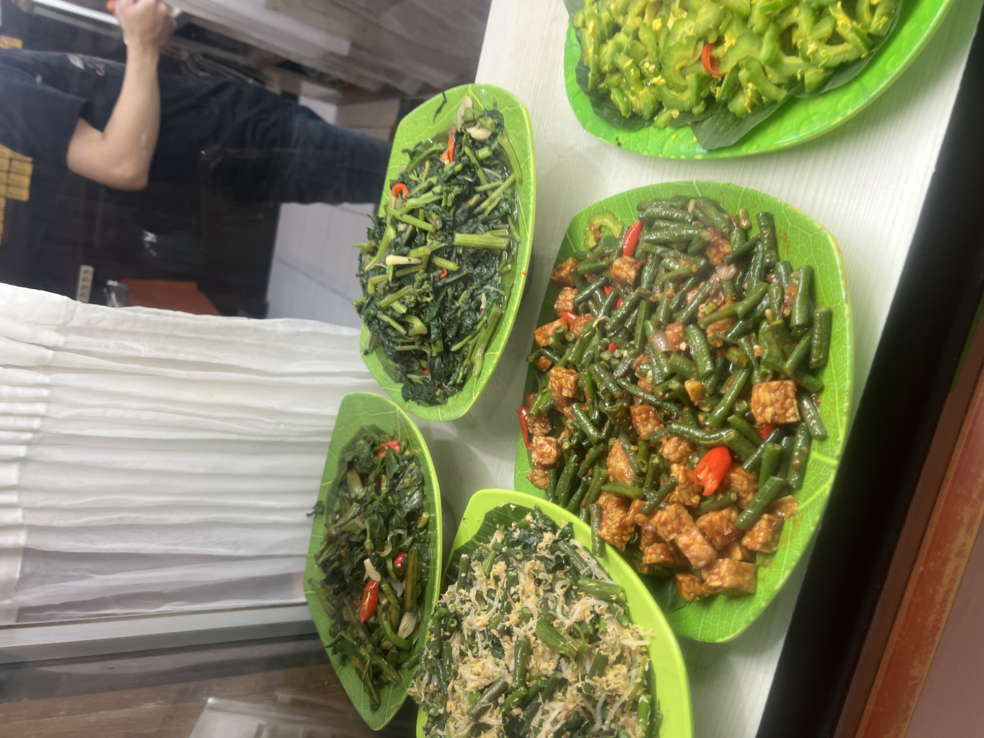 Five plates of Indonesian food are displayed on a white surface, possibly a counter or table, in a restaurant in Kuta, Indonesia. The plates are bright green and shaped like large leaves. From left to right, the first plate contains a stir-fried vegetable dish with shredded coconut and what appears to be green beans. The second plate holds a stir-fry of dark green leafy vegetables with some red chili peppers and onions. The third plate features green beans stir-fried with cubed tempeh and red chili peppers. The fourth plate is mostly obscured but appears to contain a similar green leafy vegetable dish as the second plate. The fifth plate, on the far right, contains a light green vegetable, likely bitter melon, also with some red chili peppers. In the background, a person wearing a dark t-shirt and blue jeans is visible, facing away from the camera and partially obscured by a white curtain. The environment suggests a food stall or a casual restaurant setting.