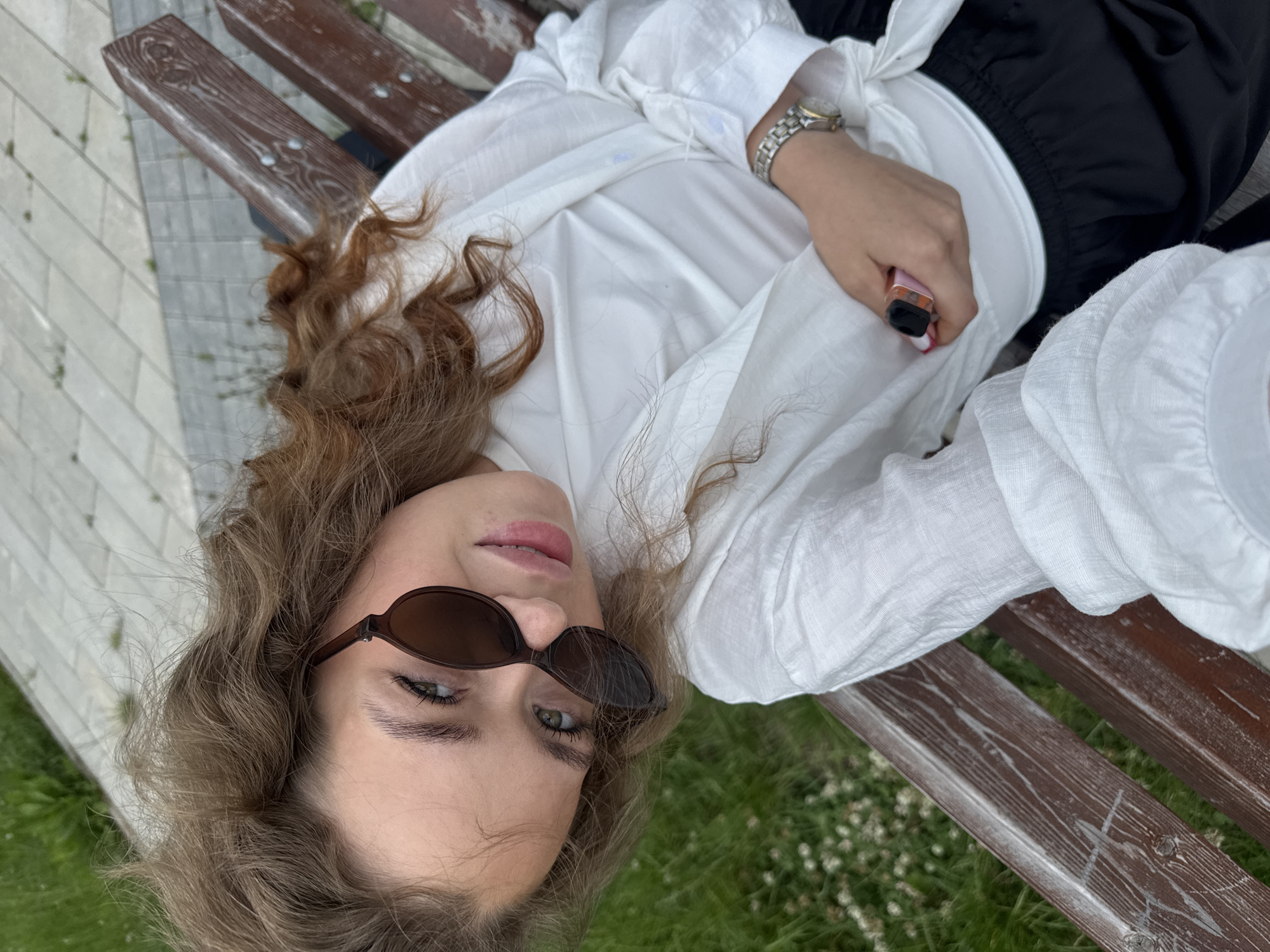 A young woman with long, wavy brown hair and green eyes, wearing sunglasses, a white button-down shirt, and black pants, lies on a wooden park bench. She is holding a pink vape device in her right hand. Her left arm rests on her stomach, and she is wearing a silver watch on her left wrist. The bench is made of dark brown wood with visible grain and some white scuff marks. The background shows a paved walkway and green grass with small white flowers. The scene appears to be outdoors in a park on a bright day. The woman’s expression is relaxed and she seems to be posing for a photo. The image was taken in Chaikovsky, Russia. There is no visible text in the image.
