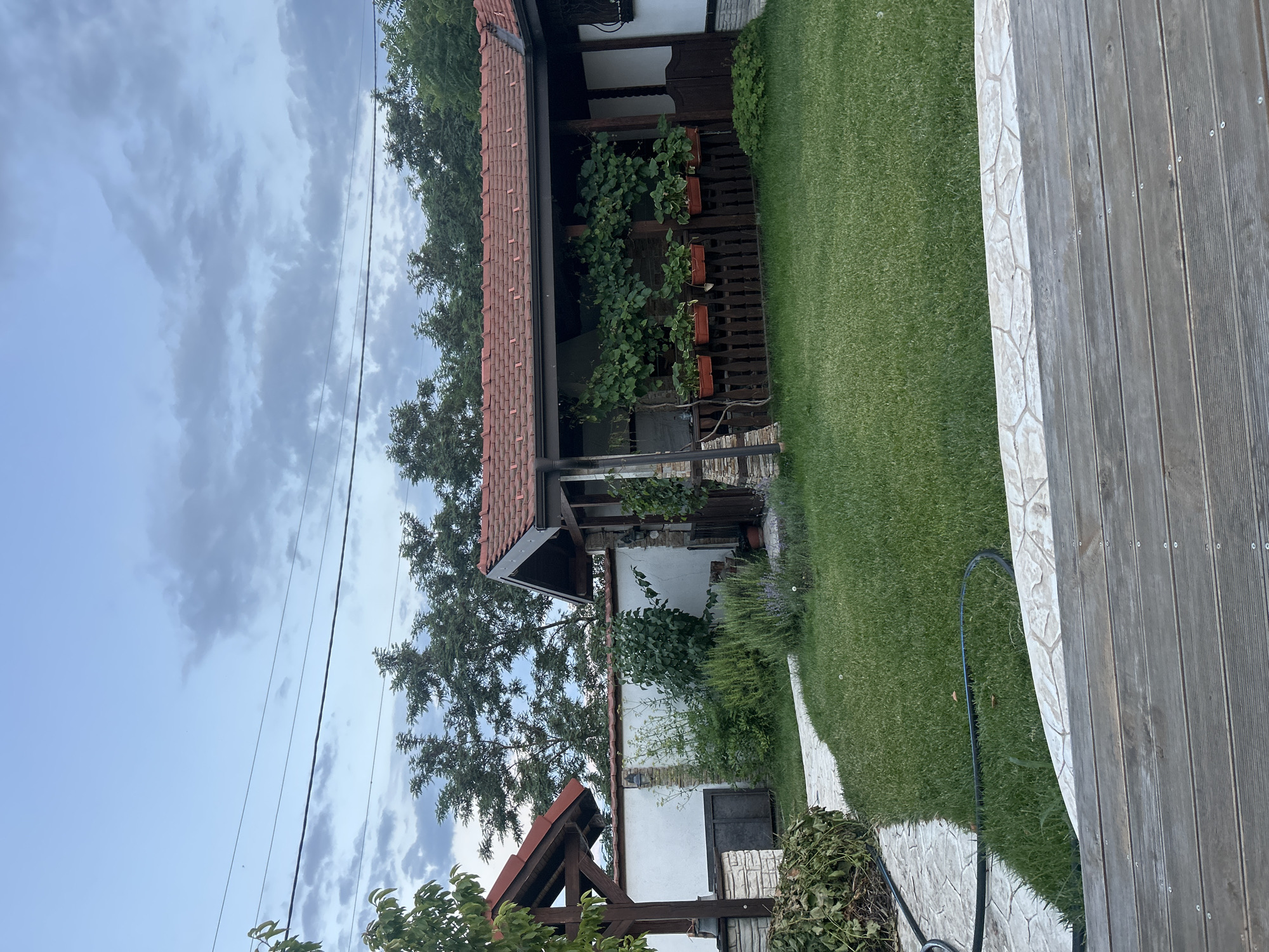A slightly overcast sky hangs over a residential property in Voyvodino, Bulgaria. The scene depicts a backyard with a well-maintained lawn, a stone pathway, and a covered patio area. The patio, with its red tile roof and wooden railings, is adorned with potted plants and climbing vines. Adjacent to the patio is a white-walled structure with a smaller covered area and a wooden support beam. Trees line the background, and power lines are visible against the sky. A wooden deck occupies the foreground, suggesting a seating or lounging area. The overall impression is one of a peaceful, lived-in outdoor space.