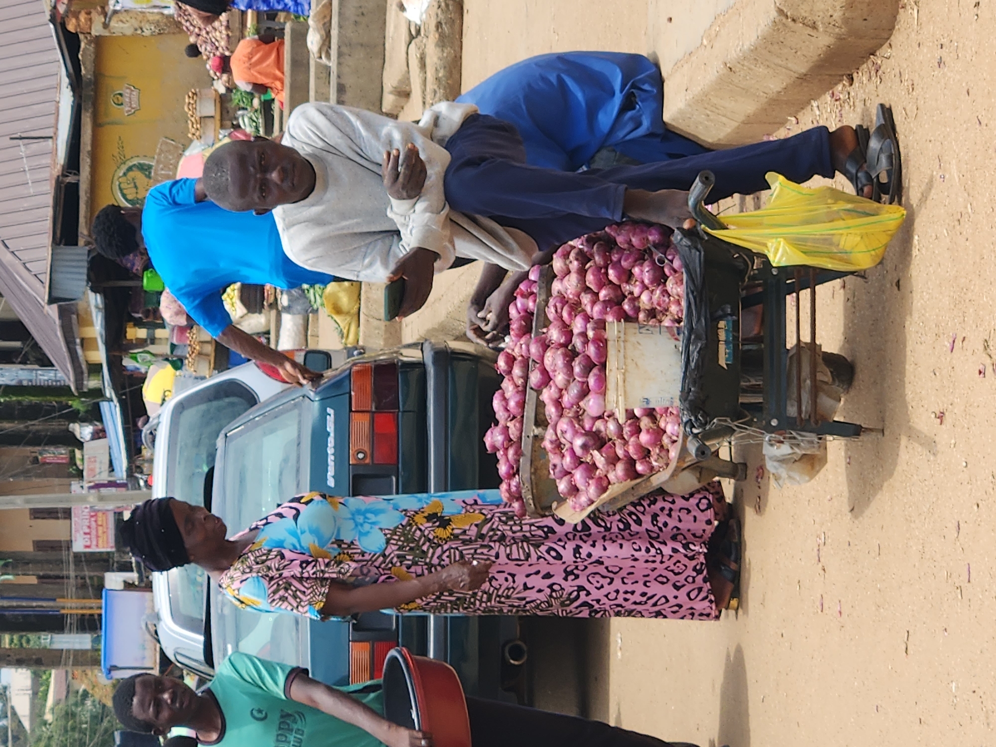 The image depicts a street market scene in Kabong, Nigeria. In the foreground, a man in a light gray long-sleeved shirt and dark blue pants sits on a concrete curb, looking at his phone. Beside him, a handcart is filled with a large quantity of red onions. A woman in a brightly colored pink, blue, and black patterned dress stands near the cart, her attention directed downwards. To the left of the woman, a young man in a green t-shirt is partially visible, holding a red bowl. In the background, various market stalls and a few vehicles are present. The ground is sandy and dusty, with small debris scattered across it. The overall atmosphere suggests a typical day at a bustling local market.