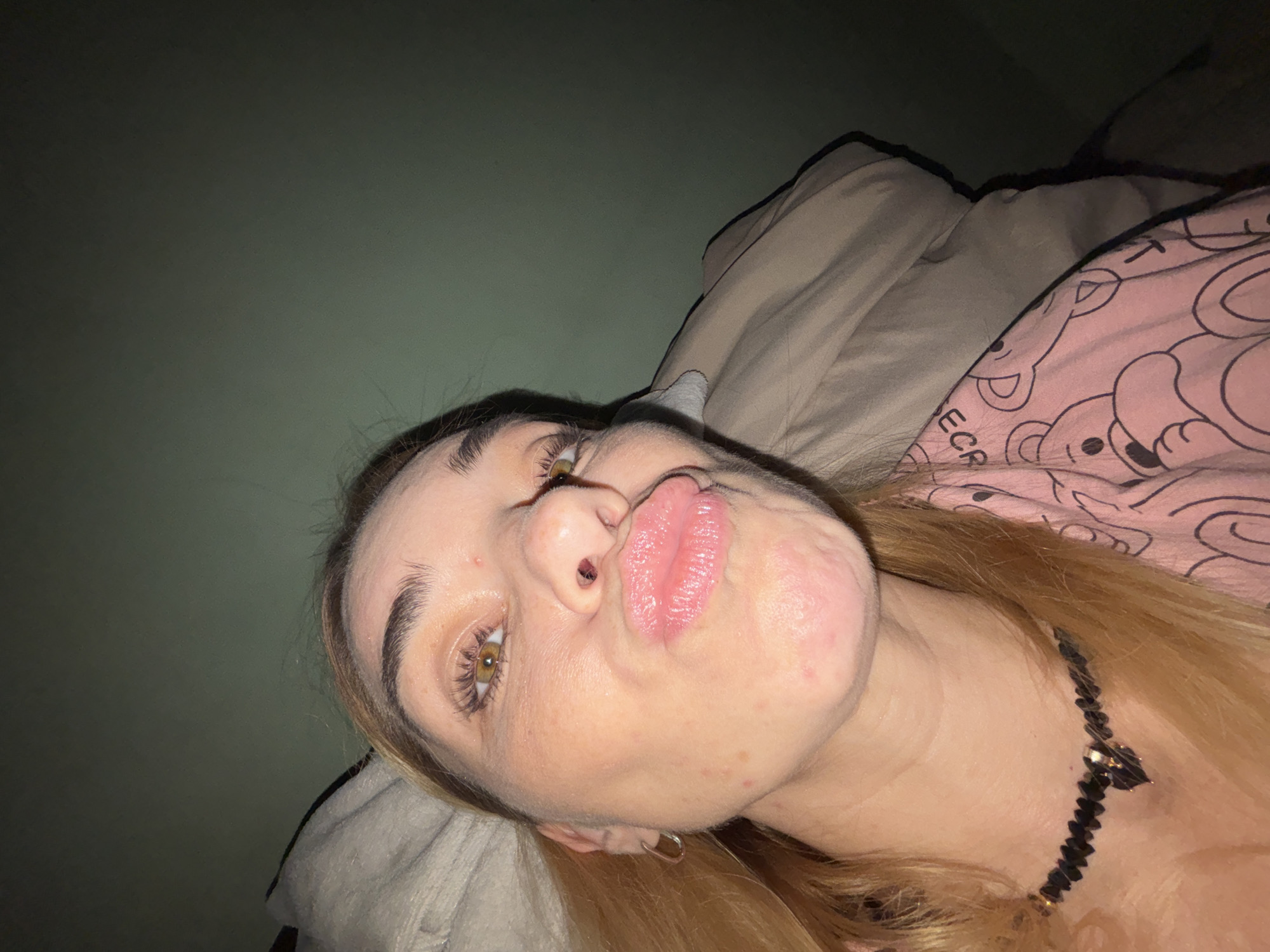An overhead, extreme close-up, selfie-style shot shows a woman with blonde hair and brown eyes puckering her lips. She is wearing a black beaded necklace with a dark pendant and a pink t-shirt with a teddy bear pattern. The background consists of a dark green wall and a rumpled beige duvet. The setting is indoors, possibly a bedroom, with no discernible details about the city of Unknown, Russia.