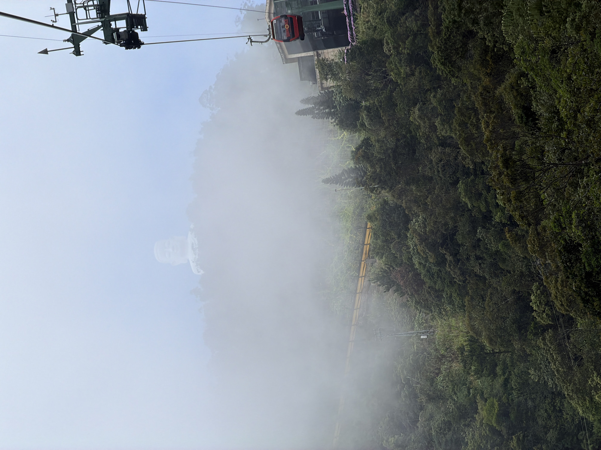 The image depicts a foggy mountain landscape with a cable car system in the foreground and a large statue visible in the distance. Specifically, this scene is from Ba Na Hills in Đà Nẵng, Vietnam. A red cable car cabin is suspended from a green metal support structure, moving towards the right side of the frame. The cabin has "SUN WORLD" written on it. Below the cable car, a lush, green mountainside is covered in dense trees and vegetation. A yellow railing is visible winding along the mountain. In the far distance, partially obscured by thick fog and clouds, the upper portion of a large white statue, likely of Buddha, can be seen emerging from the mountain peak. The sky is overcast and hazy, contributing to the misty atmosphere.