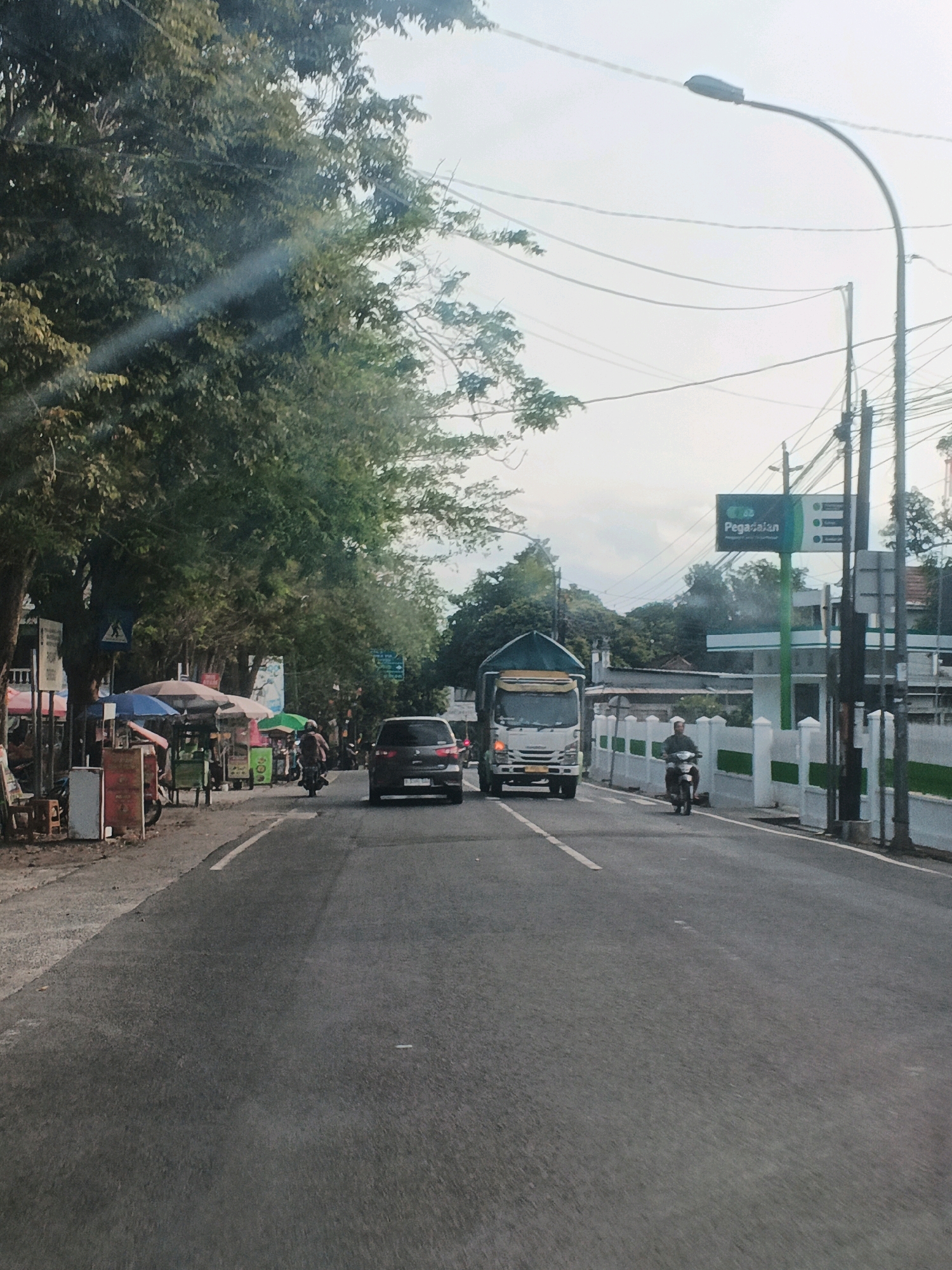 The image depicts a street scene in Minggir, Indonesia. On the right side of the road, a white and green building with a prominent "Pegadaian" sign is visible, indicating a pawnshop. A streetlight arches over the road, and numerous power lines crisscross the sky. A motorcyclist is seen riding on the right side of the road in front of the building. On the left side of the road, several vendors are set up under umbrellas, selling various goods from stalls. Trees line the left side of the road, providing shade. Traffic includes a dark-colored car in the foreground and a white truck in the middle ground. Other vehicles, including a scooter with a rider, are also present. The road is paved and marked with white lines. The sky is overcast.