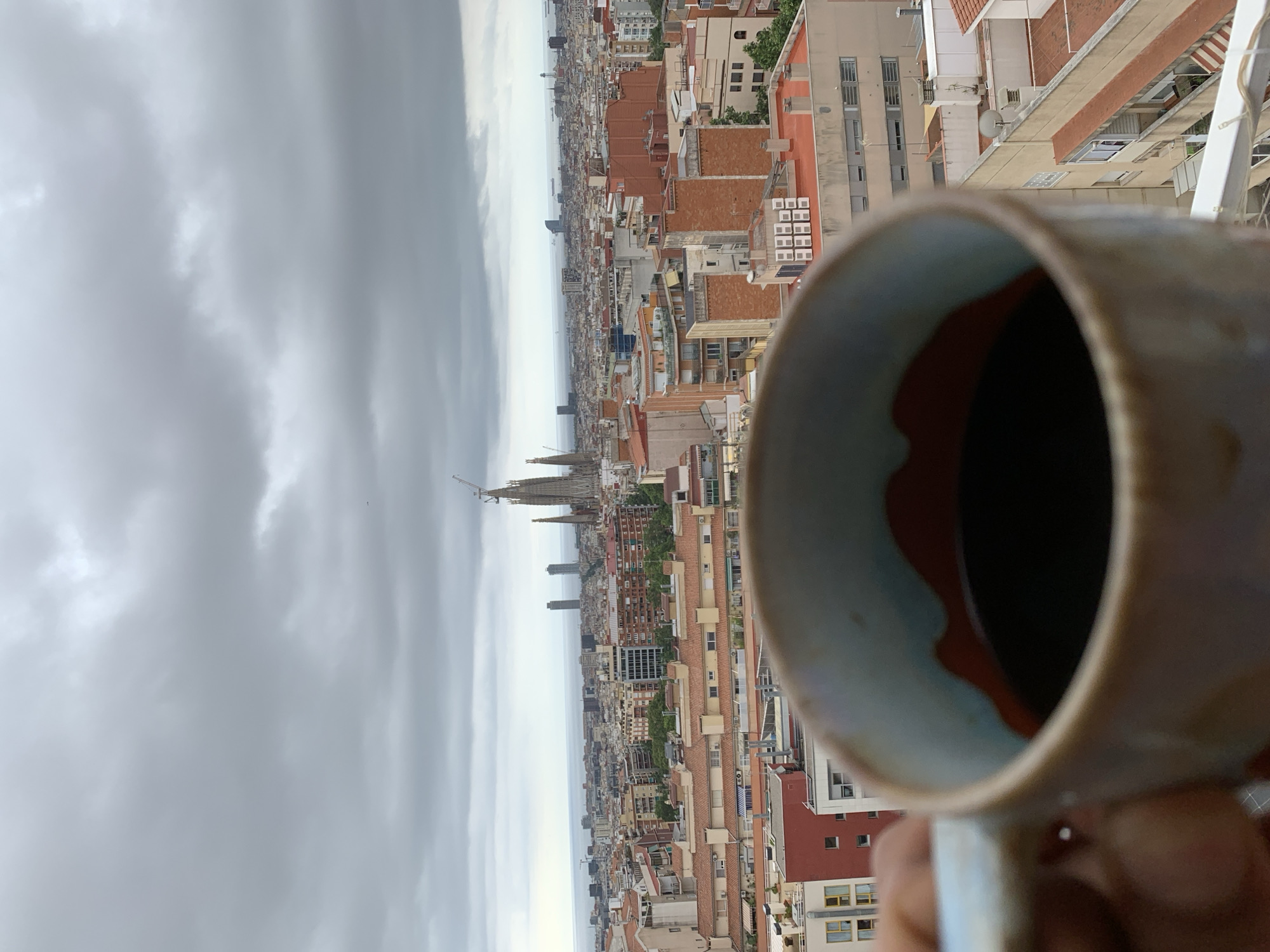 The image is a close-up shot of a mug of coffee, held by a hand, with a panoramic view of Barcelona, Spain in the background. The mug is light-colored with a blue and red interior design and is slightly out of focus, emphasizing the city view. The city is densely packed with buildings of varying heights and architectural styles, predominantly featuring terracotta tiled roofs. Dominating the skyline in the mid-ground is the Sagrada Familia, with its distinctive spires under construction, identifiable by the presence of cranes. Further in the background, the Mediterranean Sea is visible under a cloudy sky. The overall impression is a leisurely moment overlooking the cityscape.