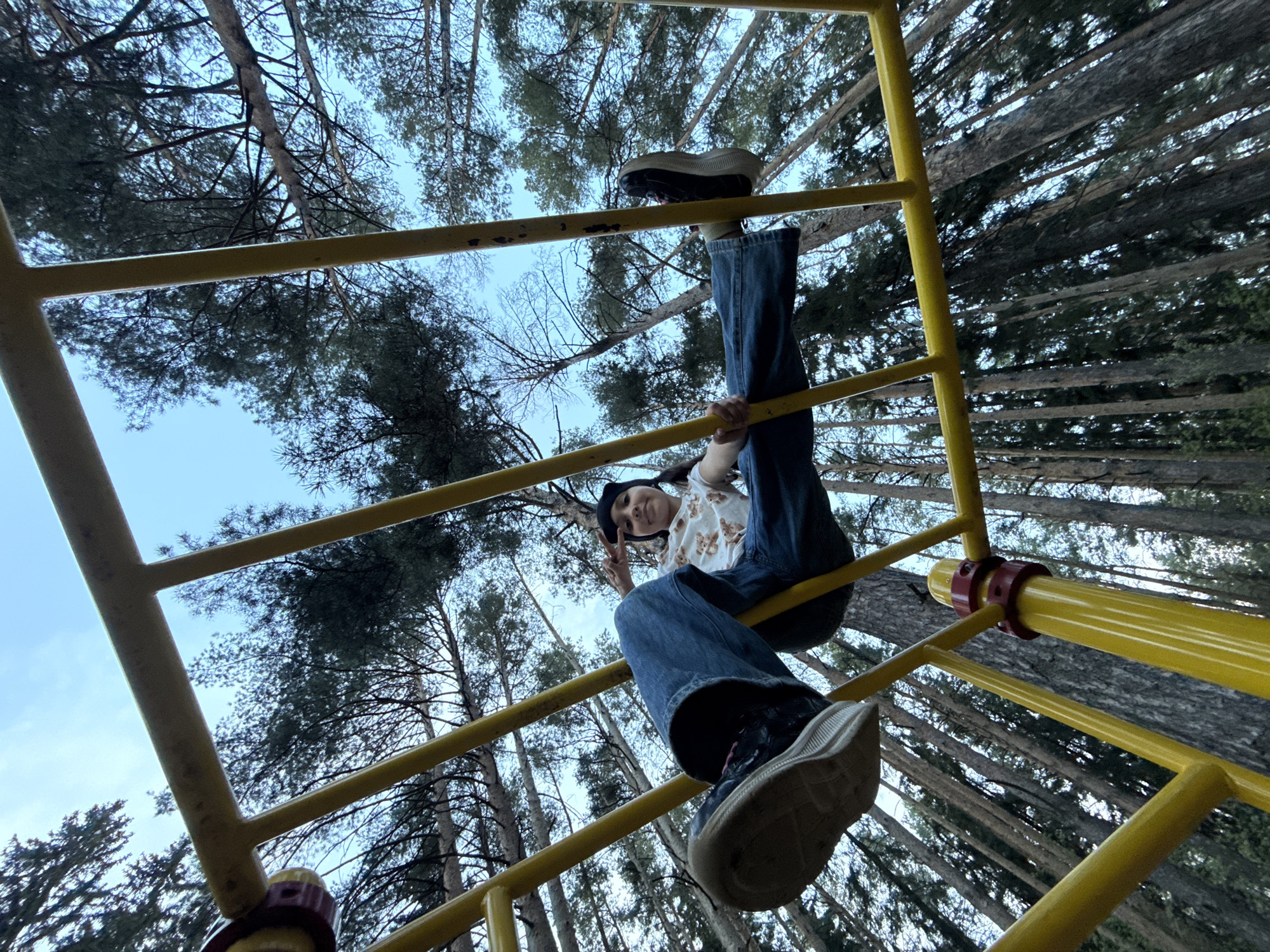 A low-angle shot depicts a person upside down on yellow monkey bars, presumably in Votkinsk, Russia. The person wears a beanie, a white shirt with a bear design, jeans, and dark sneakers. They are holding onto the bars with one hand and making a peace sign with the other. The sky and trees are visible in the background.