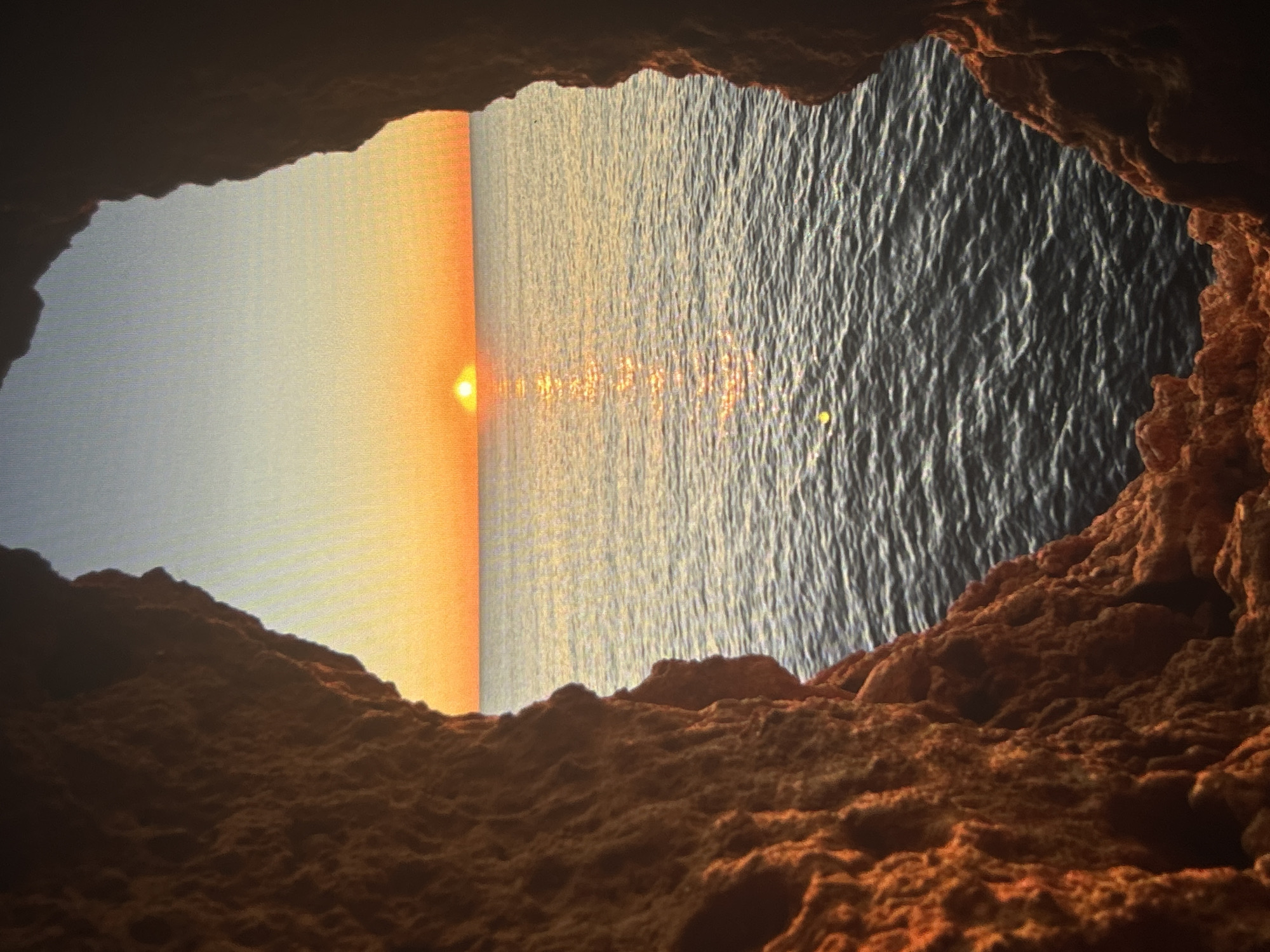 The image depicts a sunset over the ocean, viewed from inside a cave. The cave's entrance frames the scene, with dark, rocky formations in the foreground. The ocean is bisected by a gradient of light, transitioning from a dark, textured surface on the right to a bright, orange glow on the left where the sun is setting. Reflections of the sunlight shimmer on the water's surface. The scene evokes a sense of tranquility and isolation. The image cannot be located in Sofia, Bulgaria, as it depicts an ocean view.