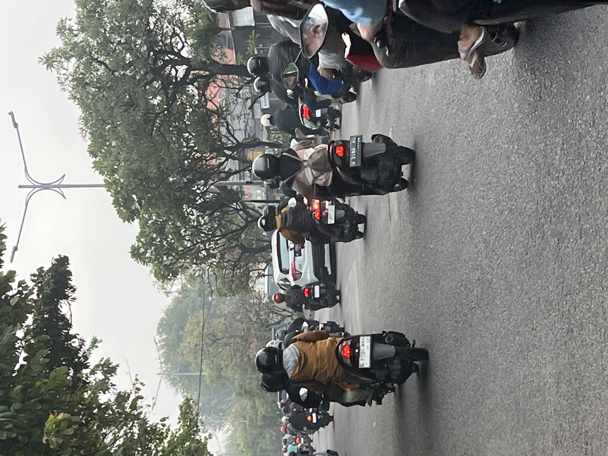 A group of motorcyclists ride on a paved road under an overcast sky. The road appears to be a city street in Sumorame, Indonesia, with trees lining the sides and buildings visible in the background. Most of the motorcyclists are wearing helmets and casual clothing, and they are riding in the same direction. Some of the motorcycles have passengers. The traffic appears to be moderate. A white car is visible in the middle of the frame, surrounded by motorcycles. The overall impression is one of a typical day with motorcycles as the primary mode of transportation.