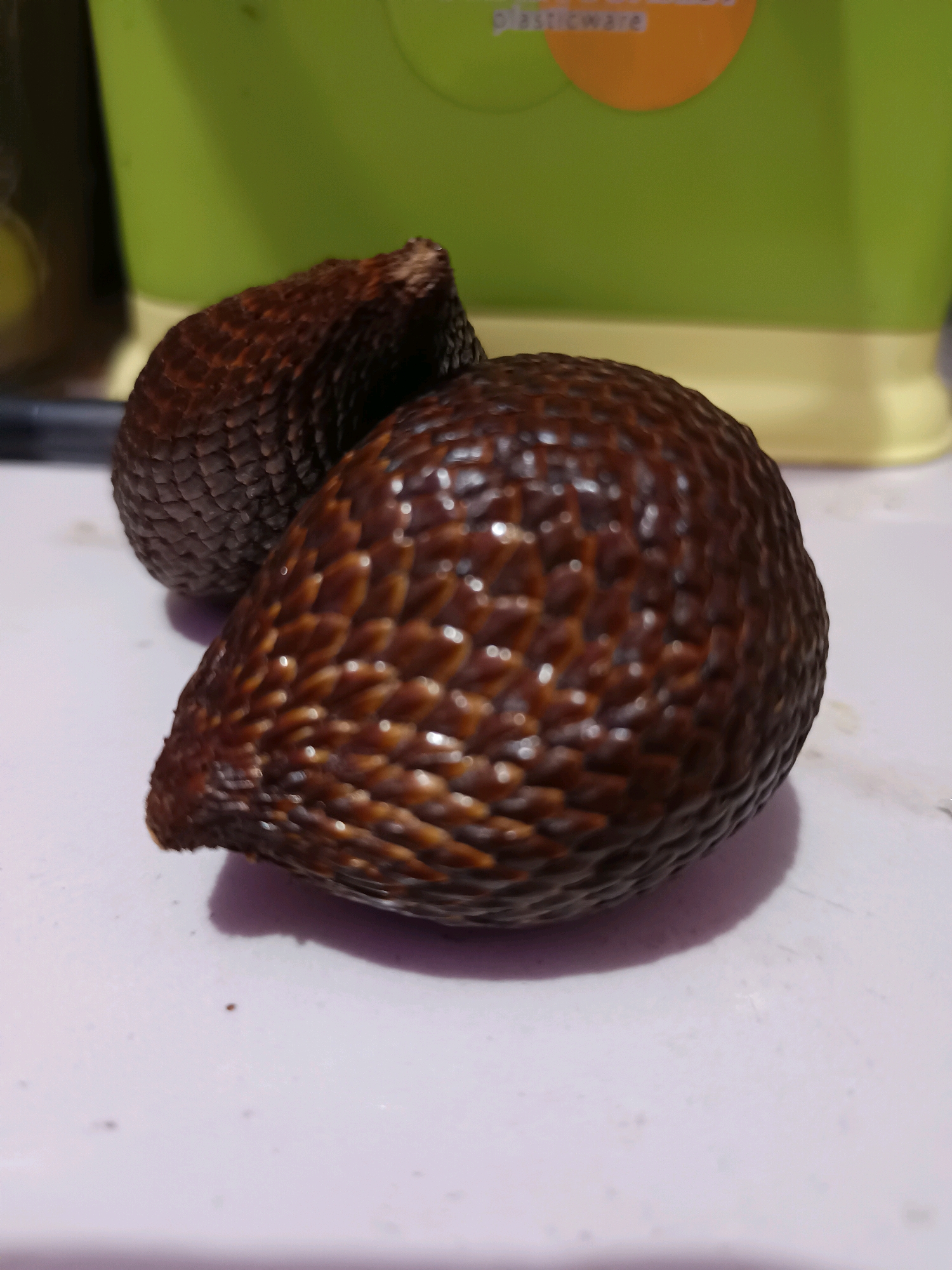 Two salak fruits are positioned on a light-colored, textured surface, likely a table. The salak fruits are dark brown with a scaly, textured skin. One fruit is closer to the foreground and appears larger, while the other is slightly behind and to the left. In the background, out of focus, is a green plastic container with a portion of an orange circle visible, suggesting it's part of a product label that reads "PLASTICWARE". The lighting suggests an indoor setting, possibly a kitchen or dining area. The location is identified as Gunung Sekar, Indonesia.